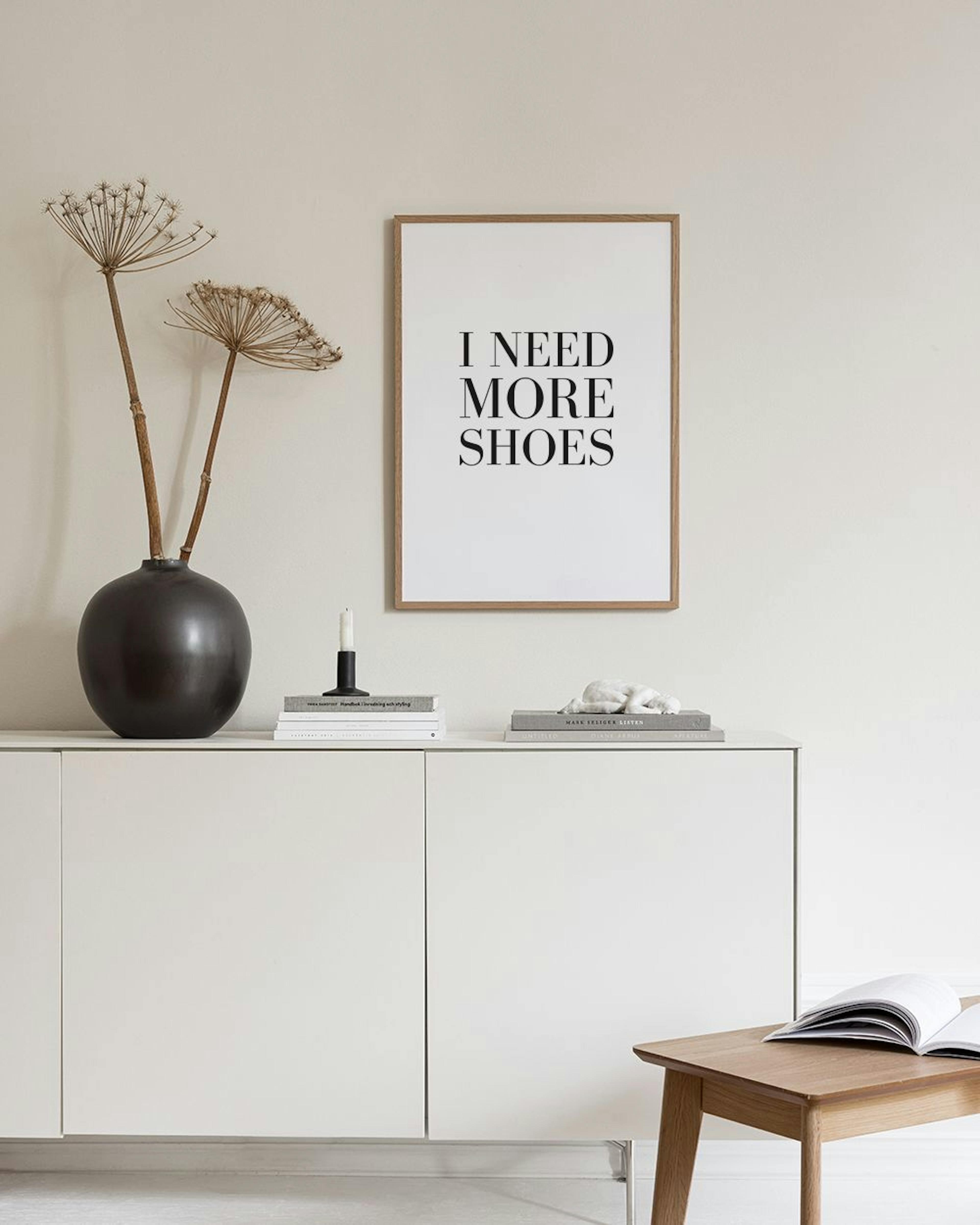 Juliste tekstillä I NEED MORE SHOES harmaata seinää vasten puupenkillä, tyynyjen, kynttilöiden ja kasvin vieressä.