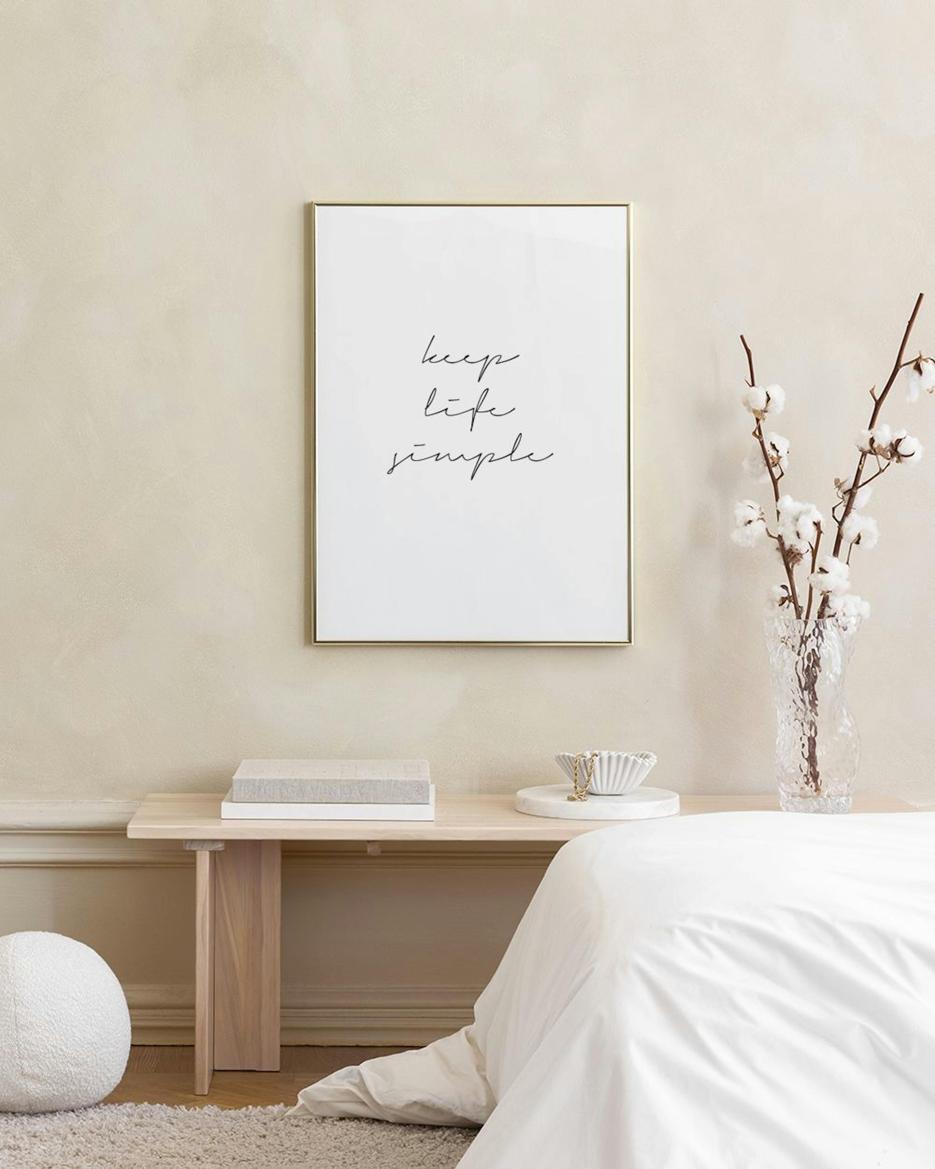 Plakát s nápisem keep life simple“ v bílém rámu, opřený o šedou zeď vedle bílé lampy a zlatých svícnů.