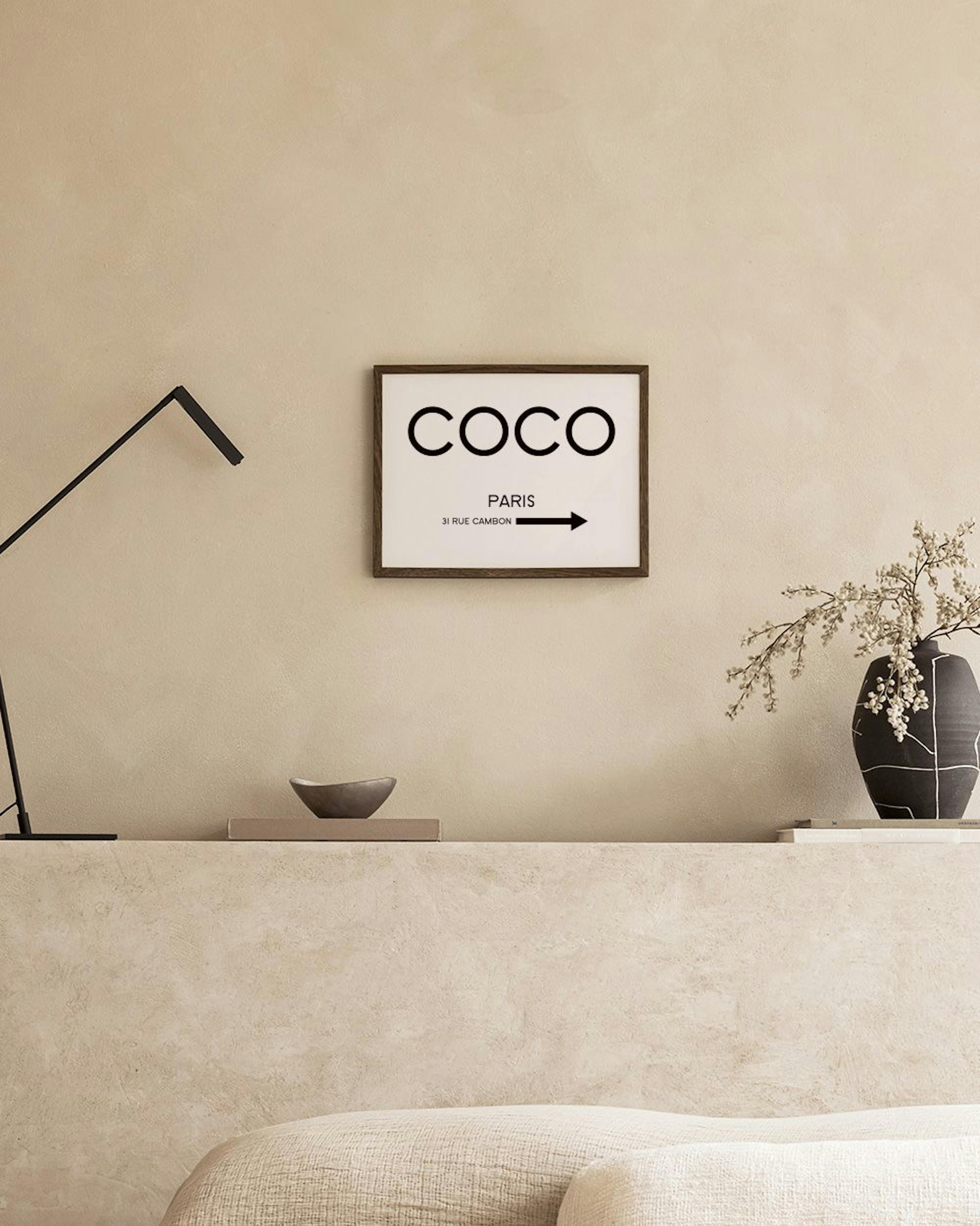 Biely plagát s čiernym nápisom COCO PARIS a šípkou visí v miestnosti nad čiernym stolíkom s knihami a vázou.