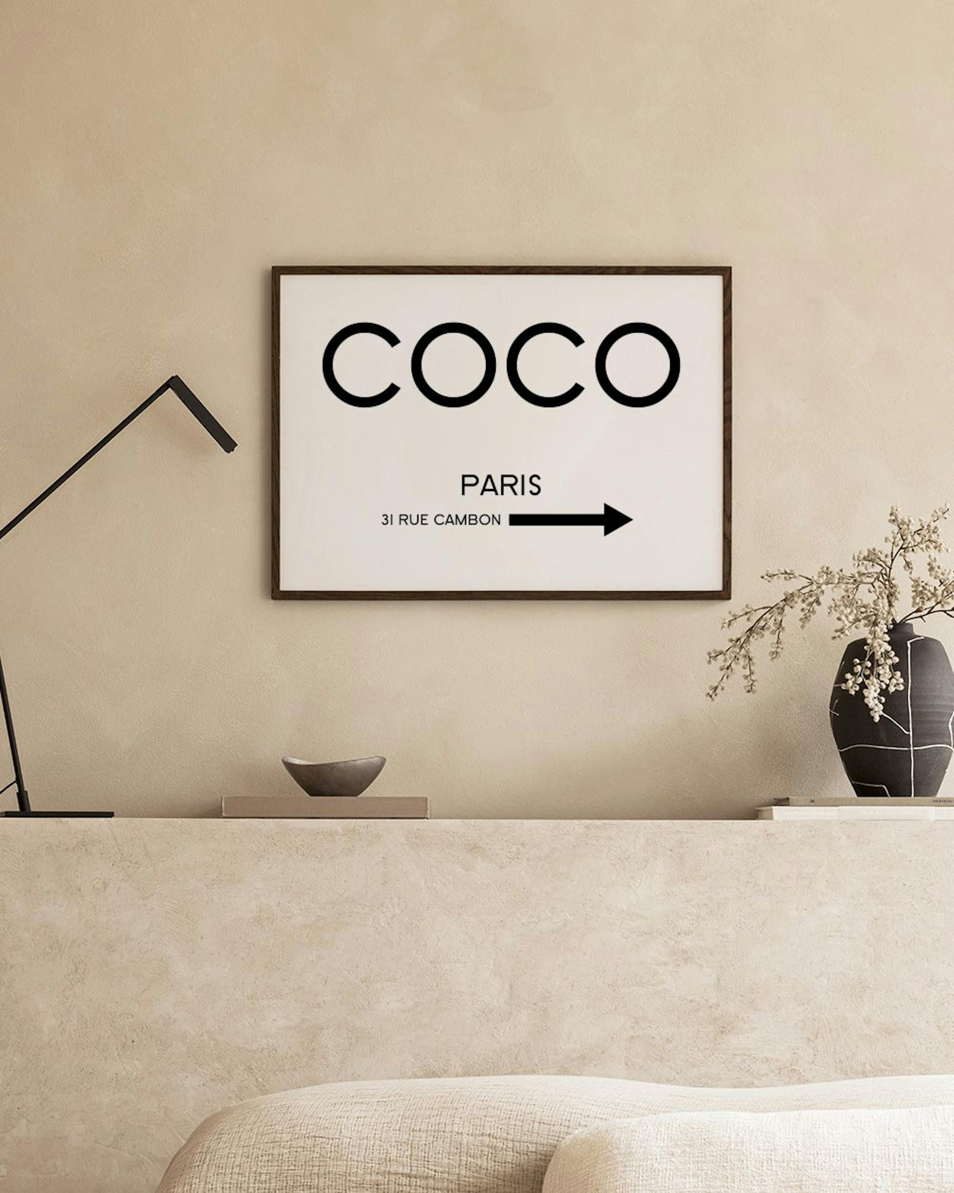 Póster blanco con texto negro COCO PARIS 31 RUE CAMBON y una flecha, sobre una pared gris oscuro con una mesa y una canasta.