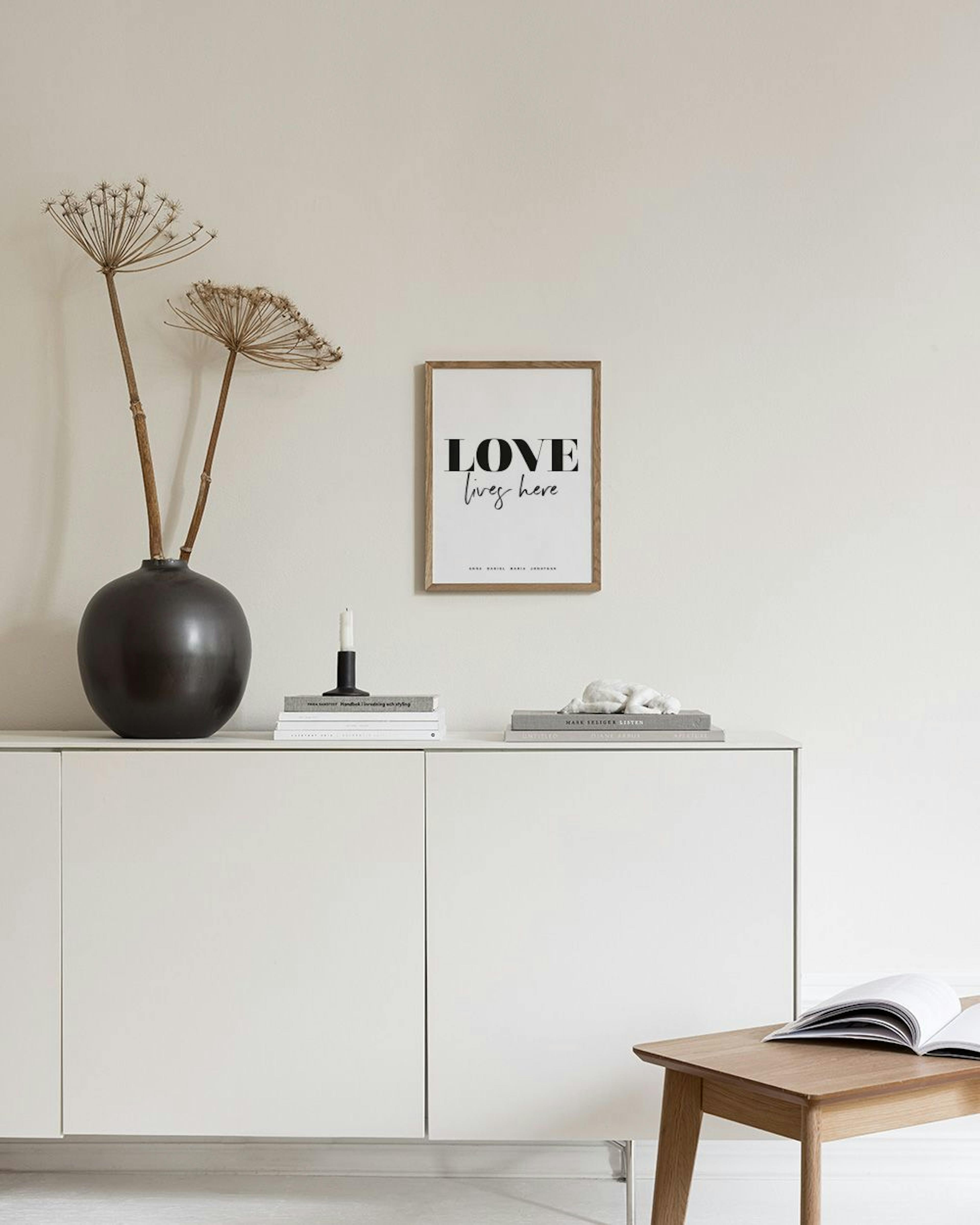 Póster personalizado con la frase LOVE lives here y nombres de familia, colgado en una pared lila de una sala moderna.