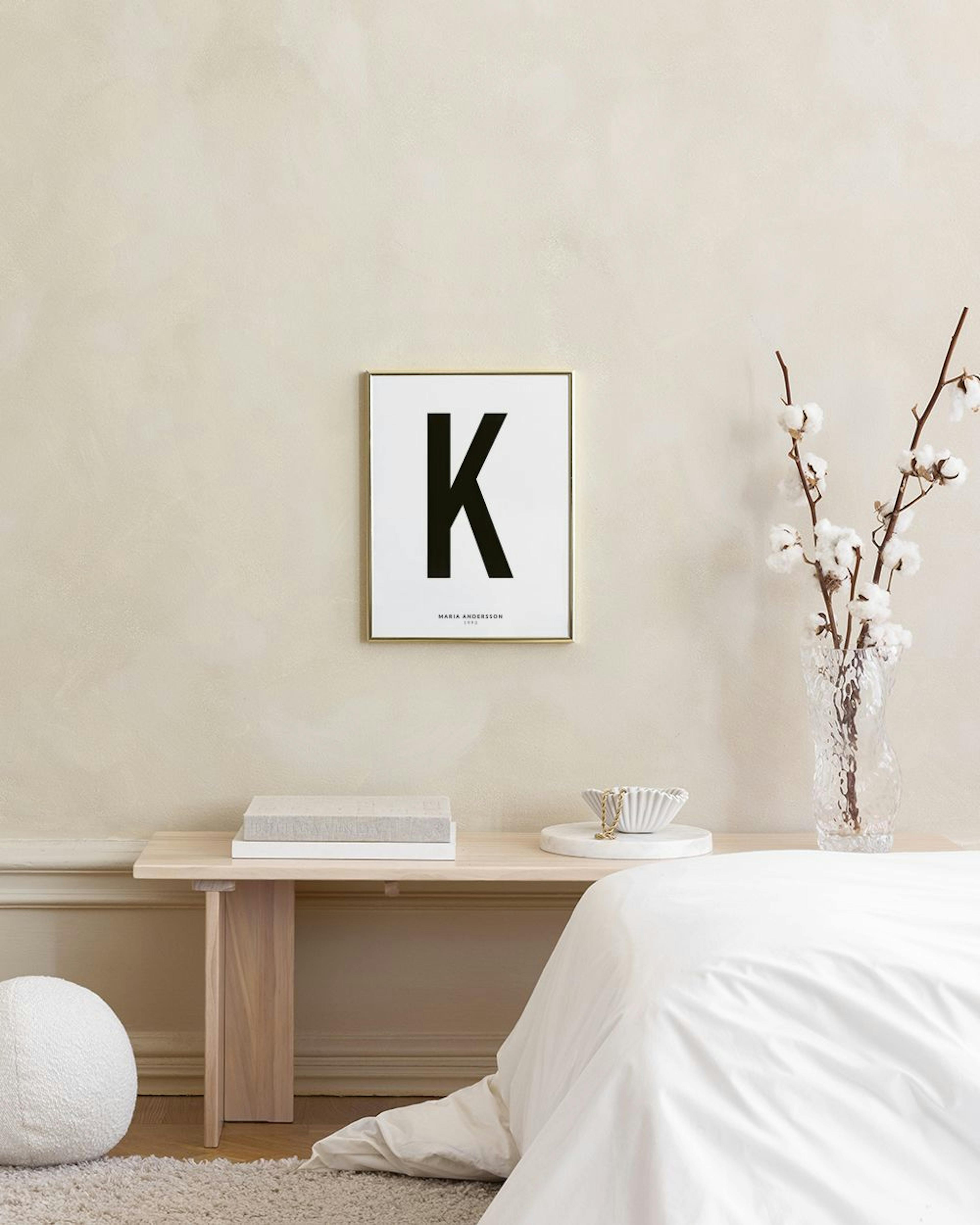 Personlig plakat med sort bogstav K og navn, hængende over en flettet bænk med keramikvaser og bøger.