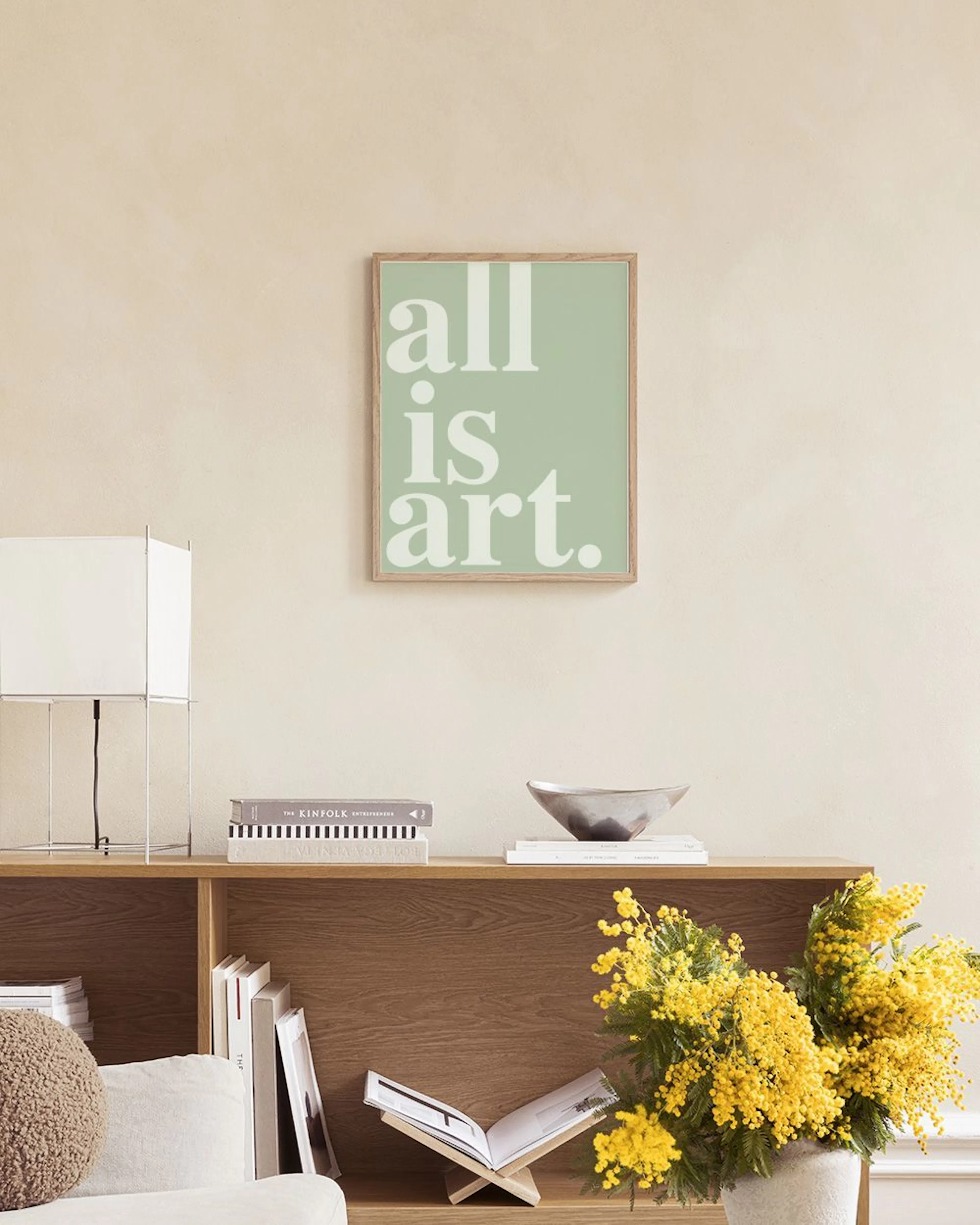 Poster med texten all is art på mintgrön bakgrund, hängande ovanför en mintgrön soffa med syrener i vas.