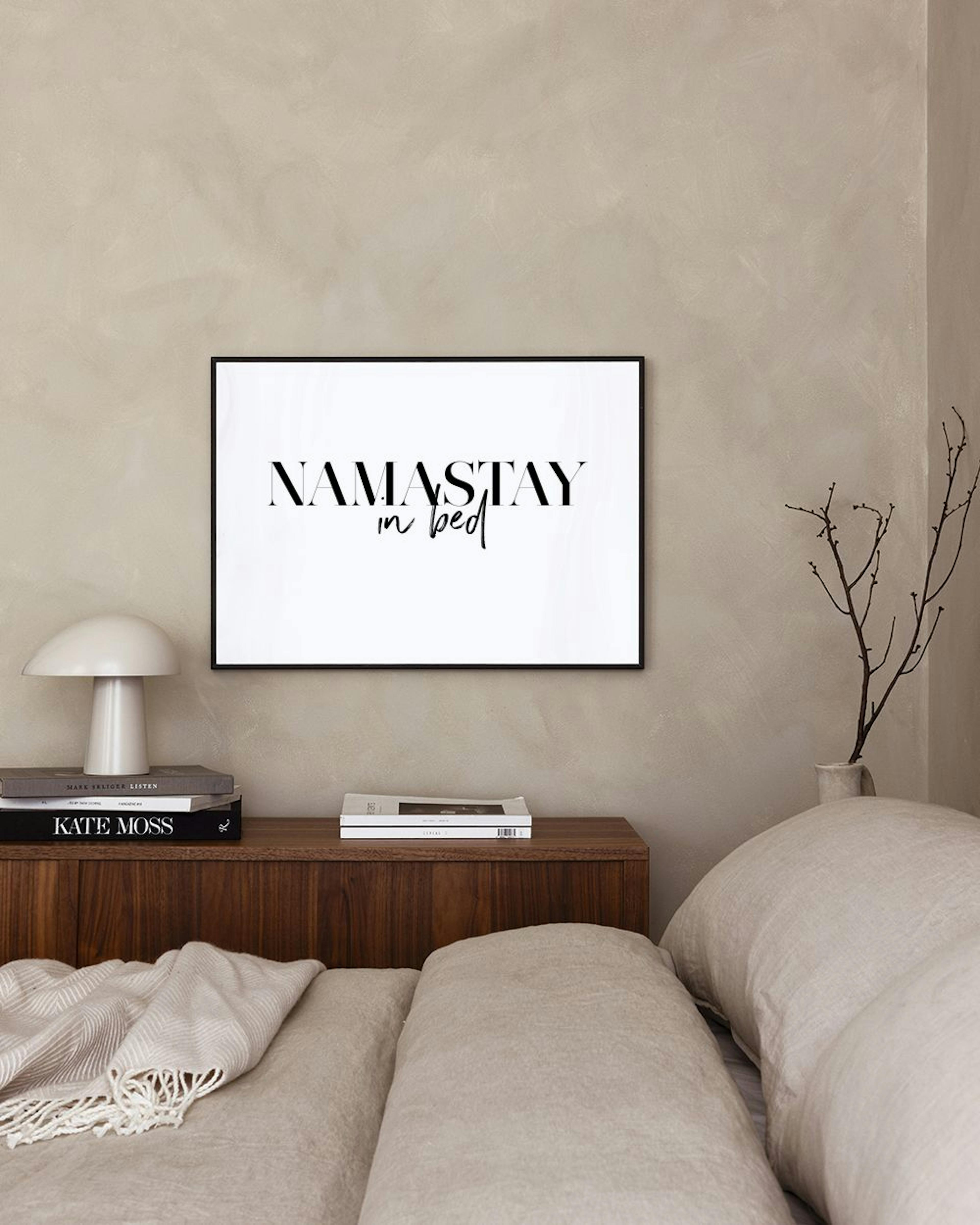 A minimalist typography print with NAMASTAY in bed in black text, hanging above a bed with gray and gold pillows.