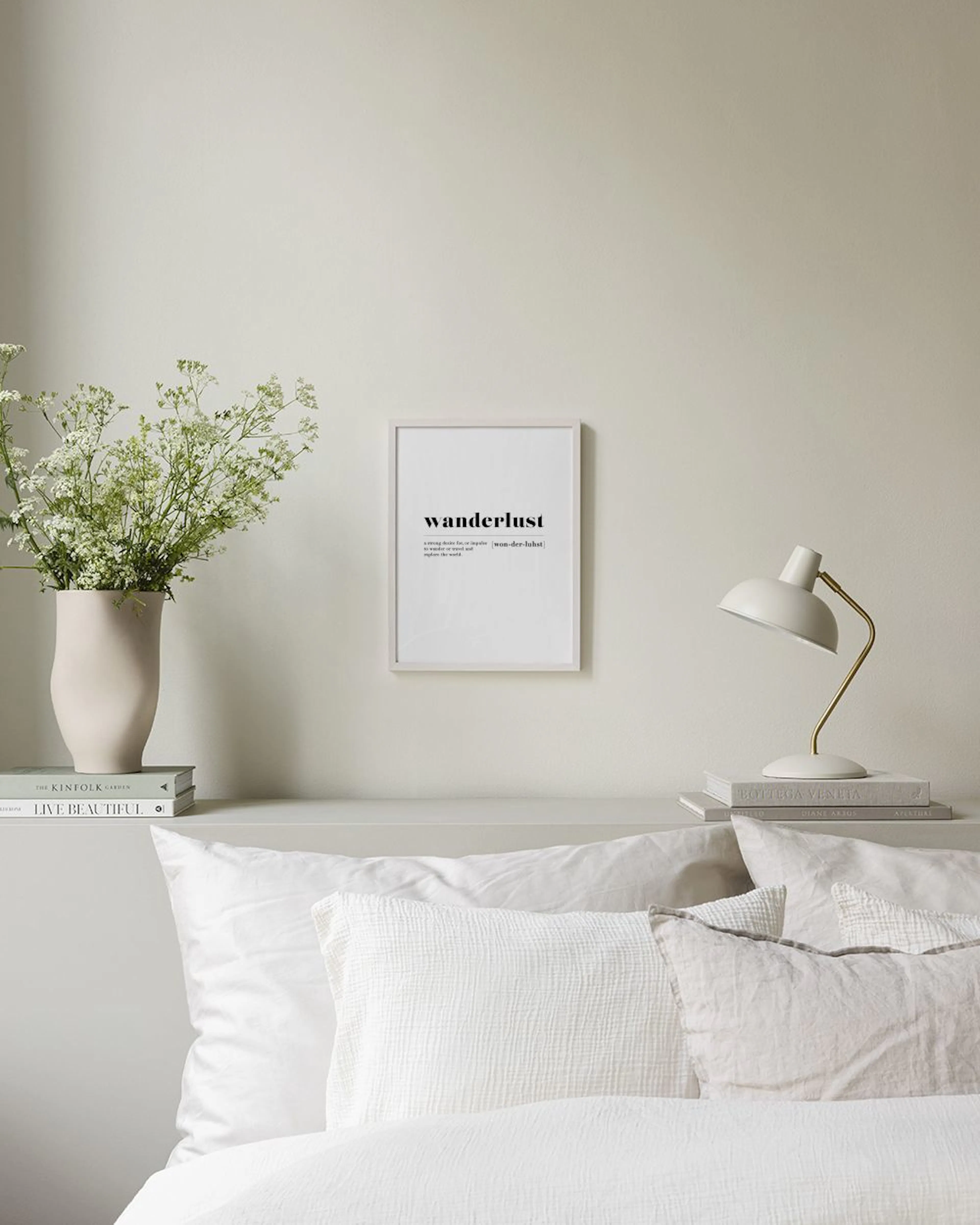 A print with the definition of wanderlust in black text on a white background, hanging above a white console table with books an
