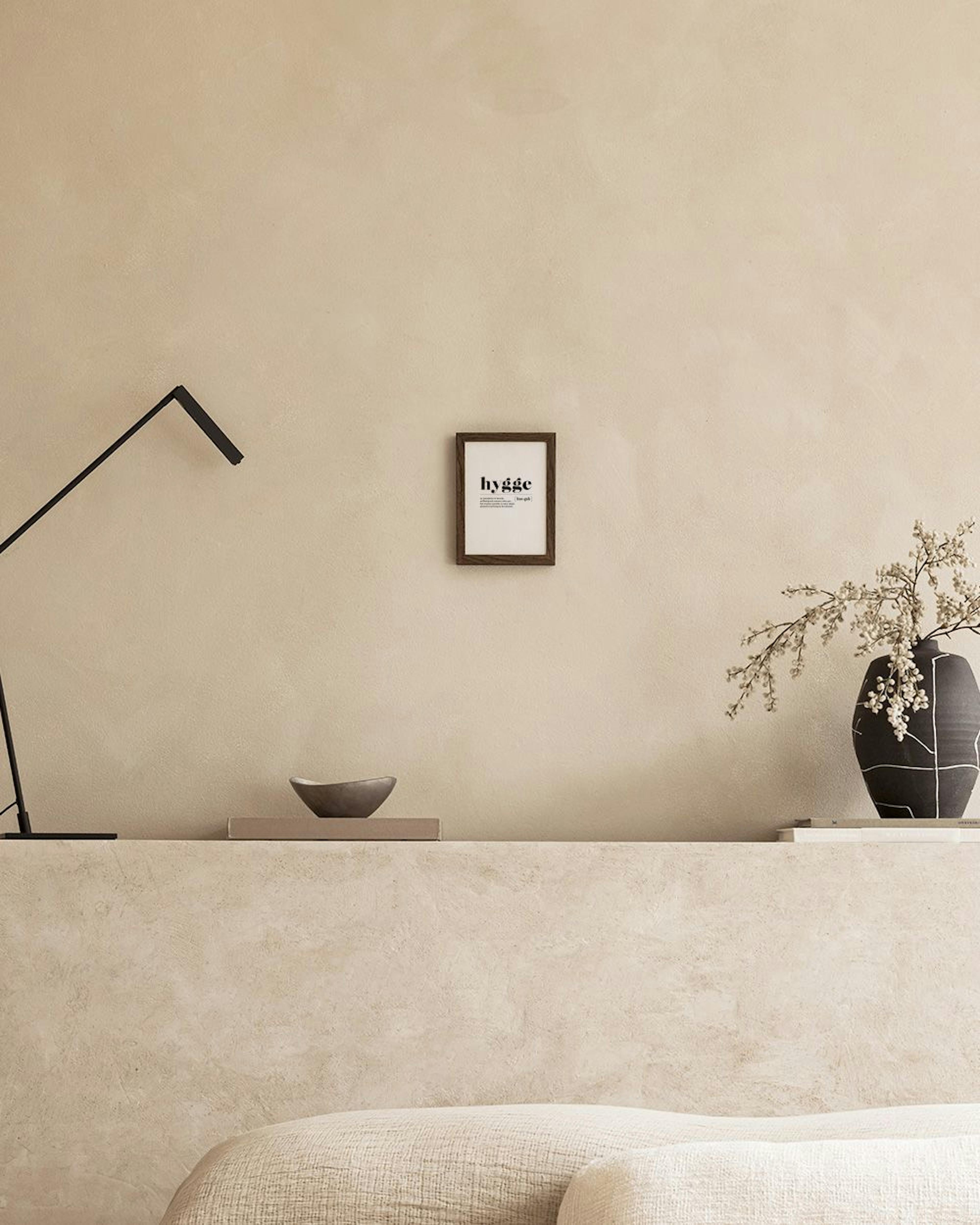 Poster minimalista com a palavra hygge e sua definição, pendurado acima de uma mesa de cabeceira com um vaso de flores e livros,
