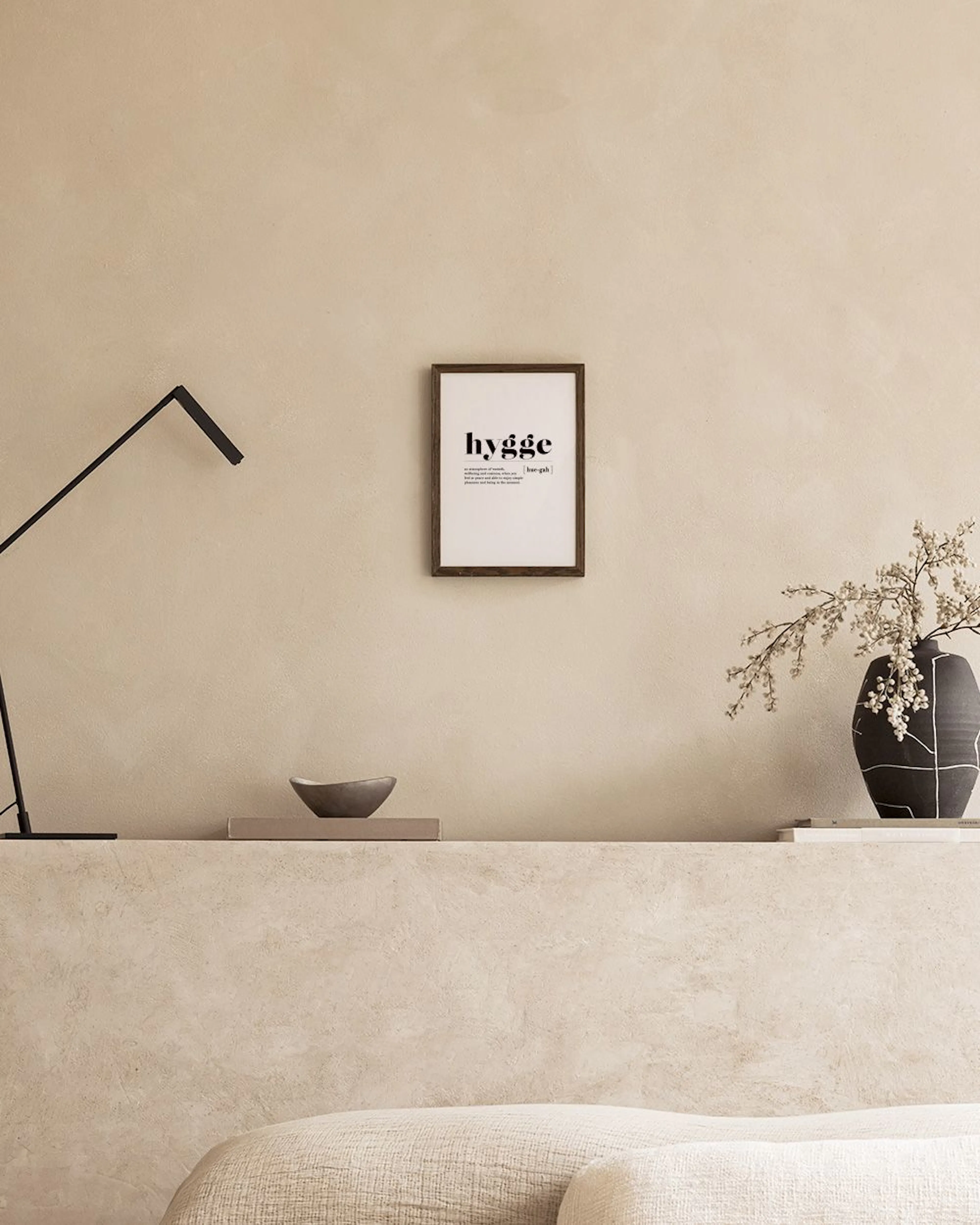 A print with the word hygge and its definition, hanging above a bedside table with a vase of flowers and books.