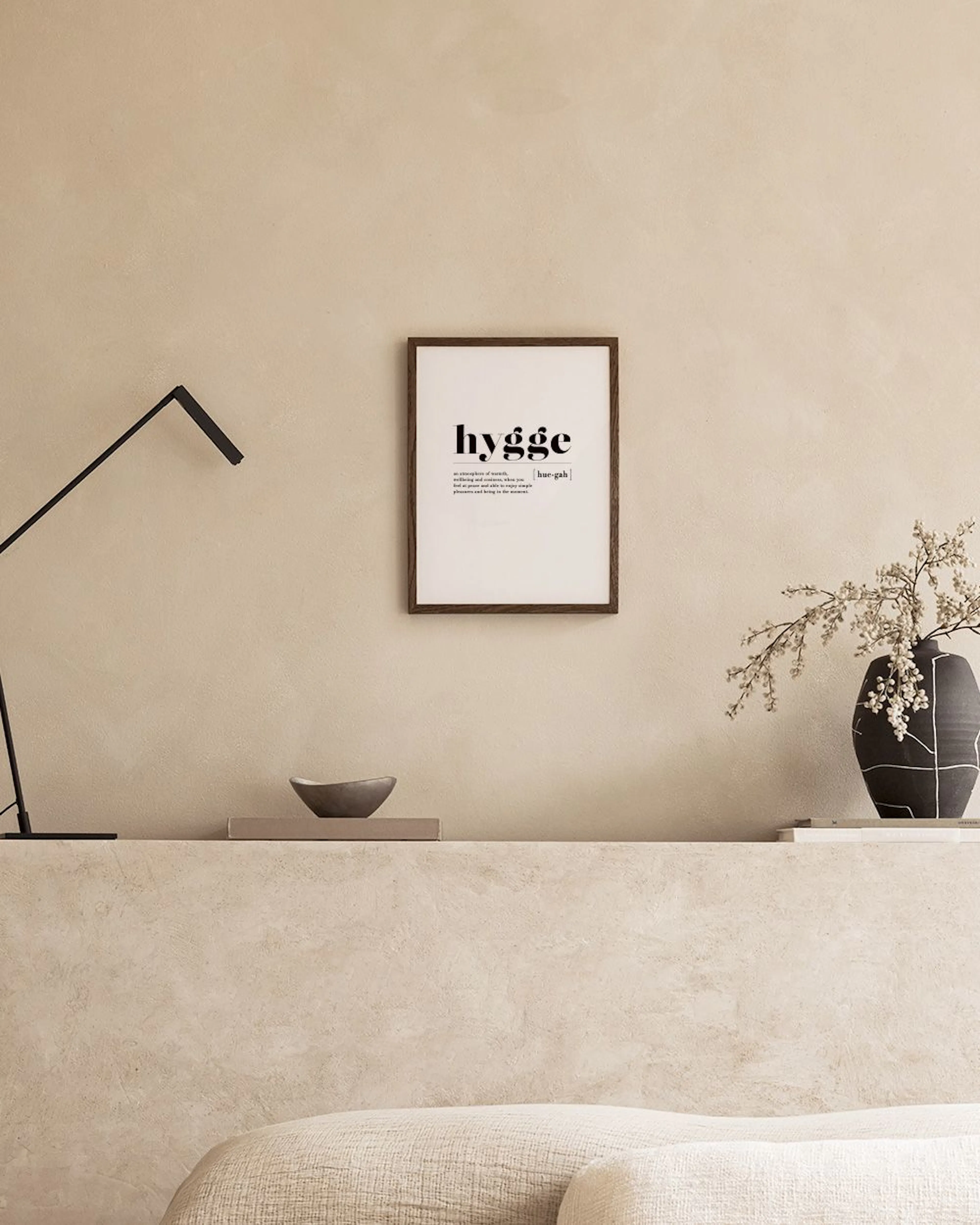 Poster Hygge mit Definition, über einem Nachttisch mit Vase und Buch in einem Schlafzimmer.