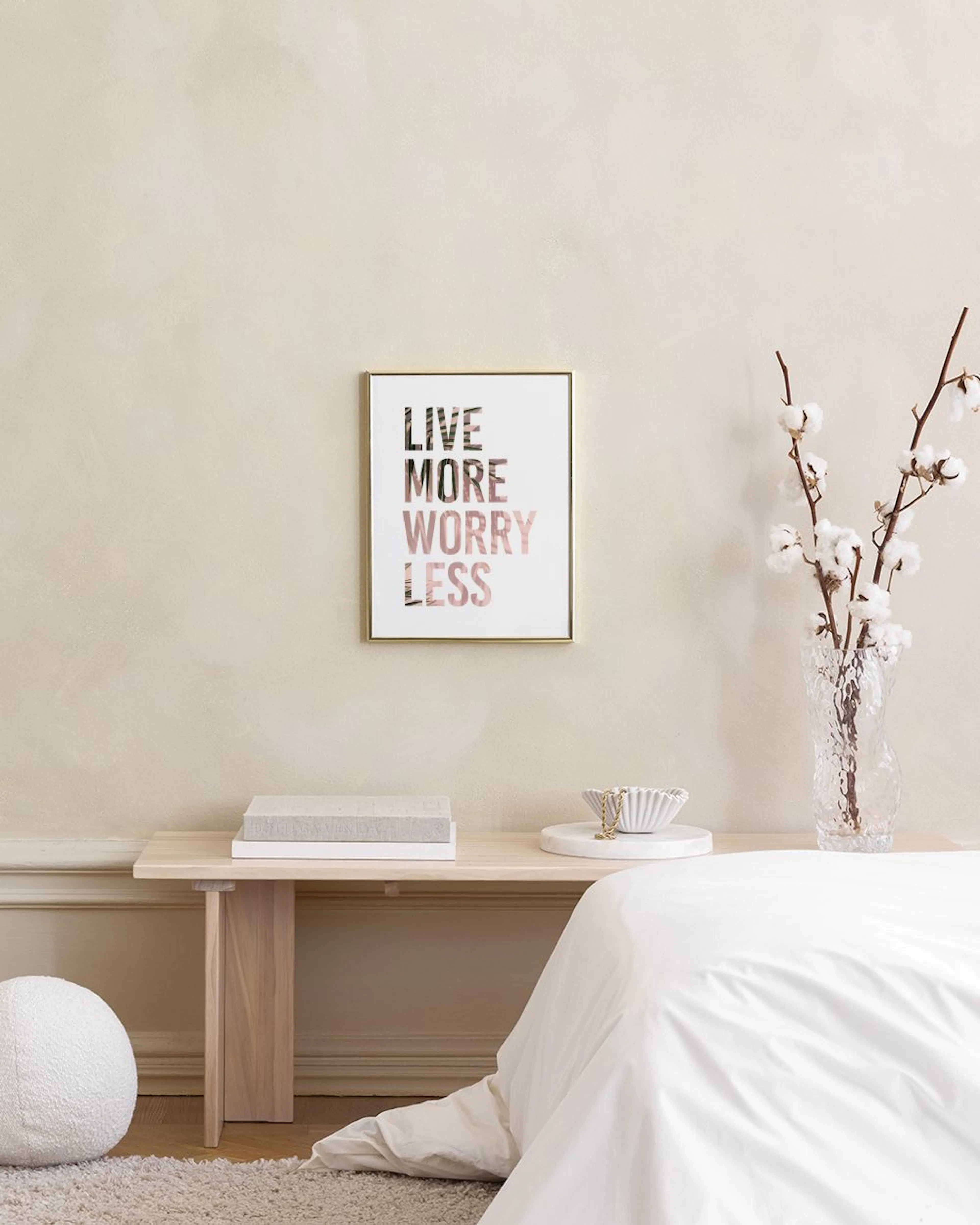 A print with the words LIVE MORE WORRY LESS in large, bold, rose gold-toned letters, hanging on a grey wall above a wooden table