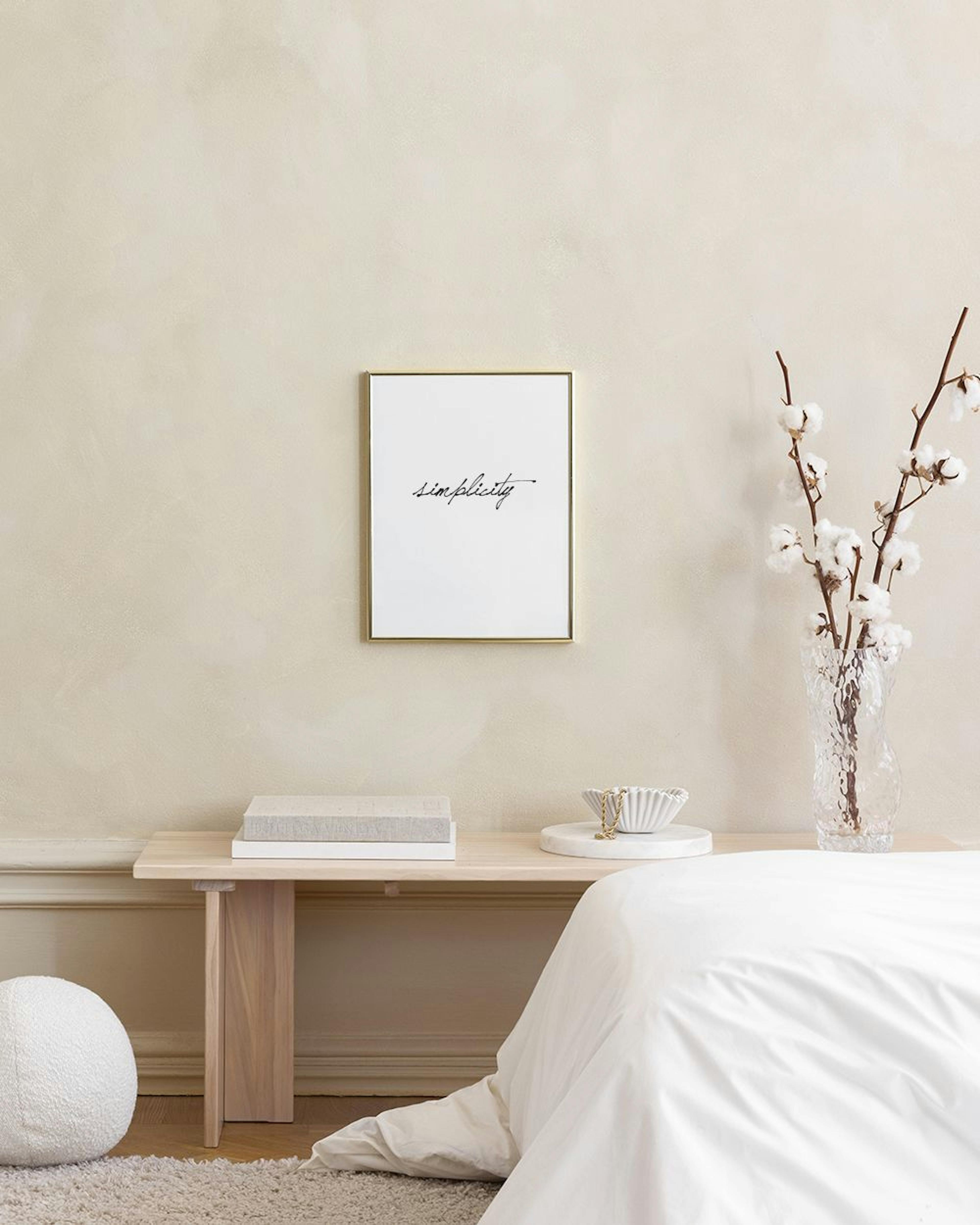 Valkoinen Simplicity-juliste nojaa vaaleanpunaisen seinän edessä pöydällä, jonka päällä on myös puinen käsi, musta rasia ja kakt