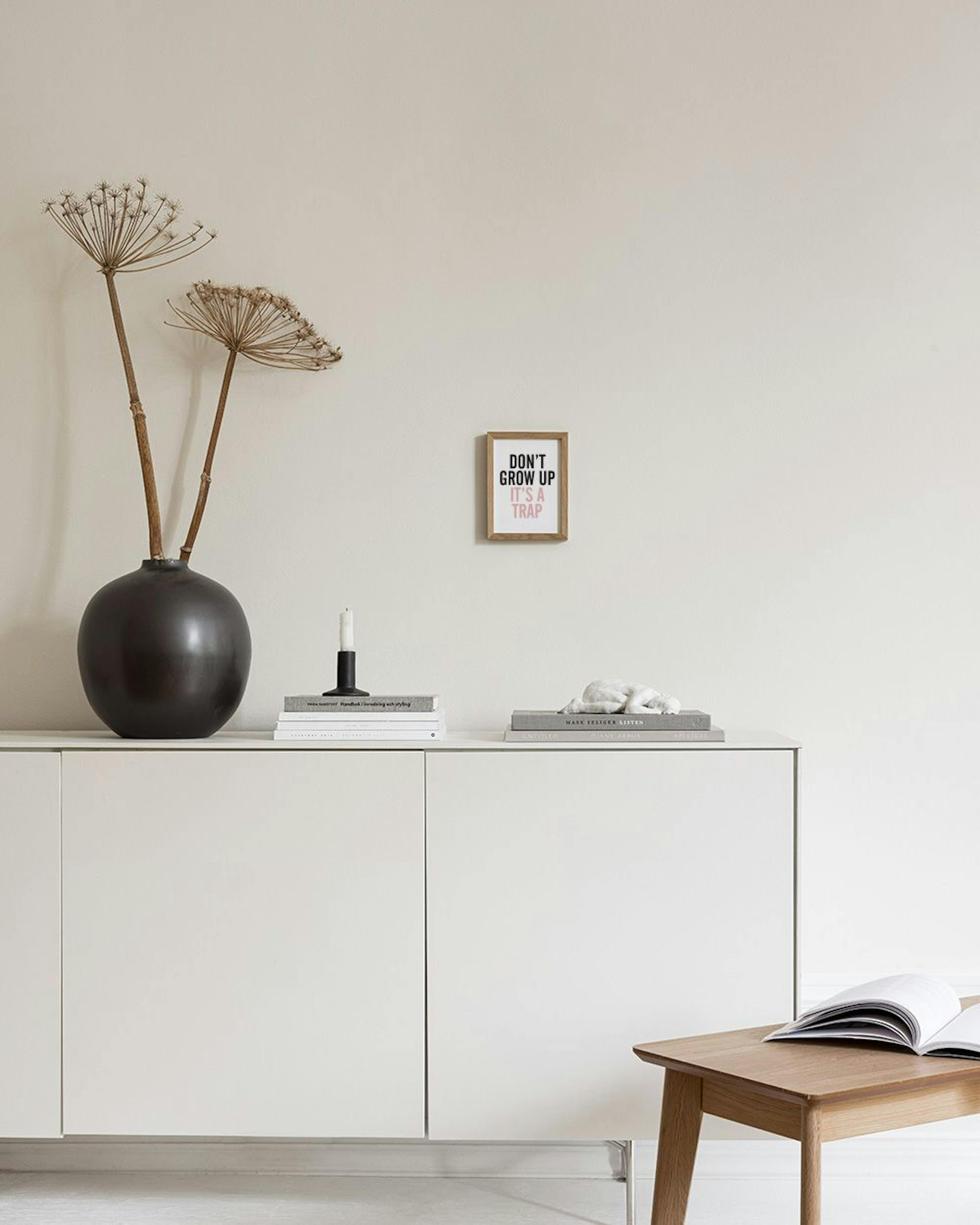 En plakat med teksten DONT GROW UP ITS A TRAP i sort og lyserød, stående på et hvidt bord ved siden af en hvid lampe og gyldne l
