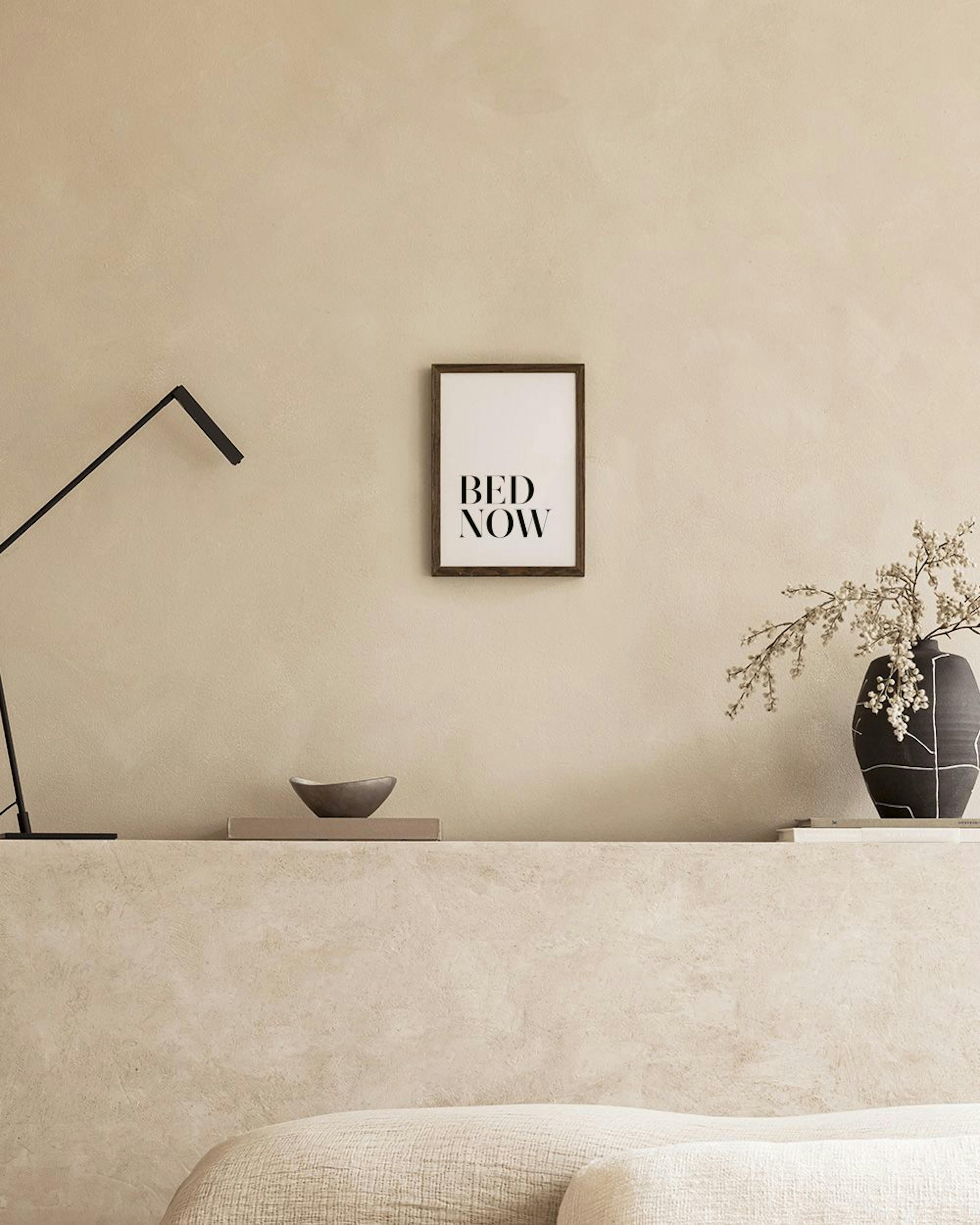 A black and white BED NOW quote print hanging above a white bedside table with a vase of flowers, next to a bed with grey beddin