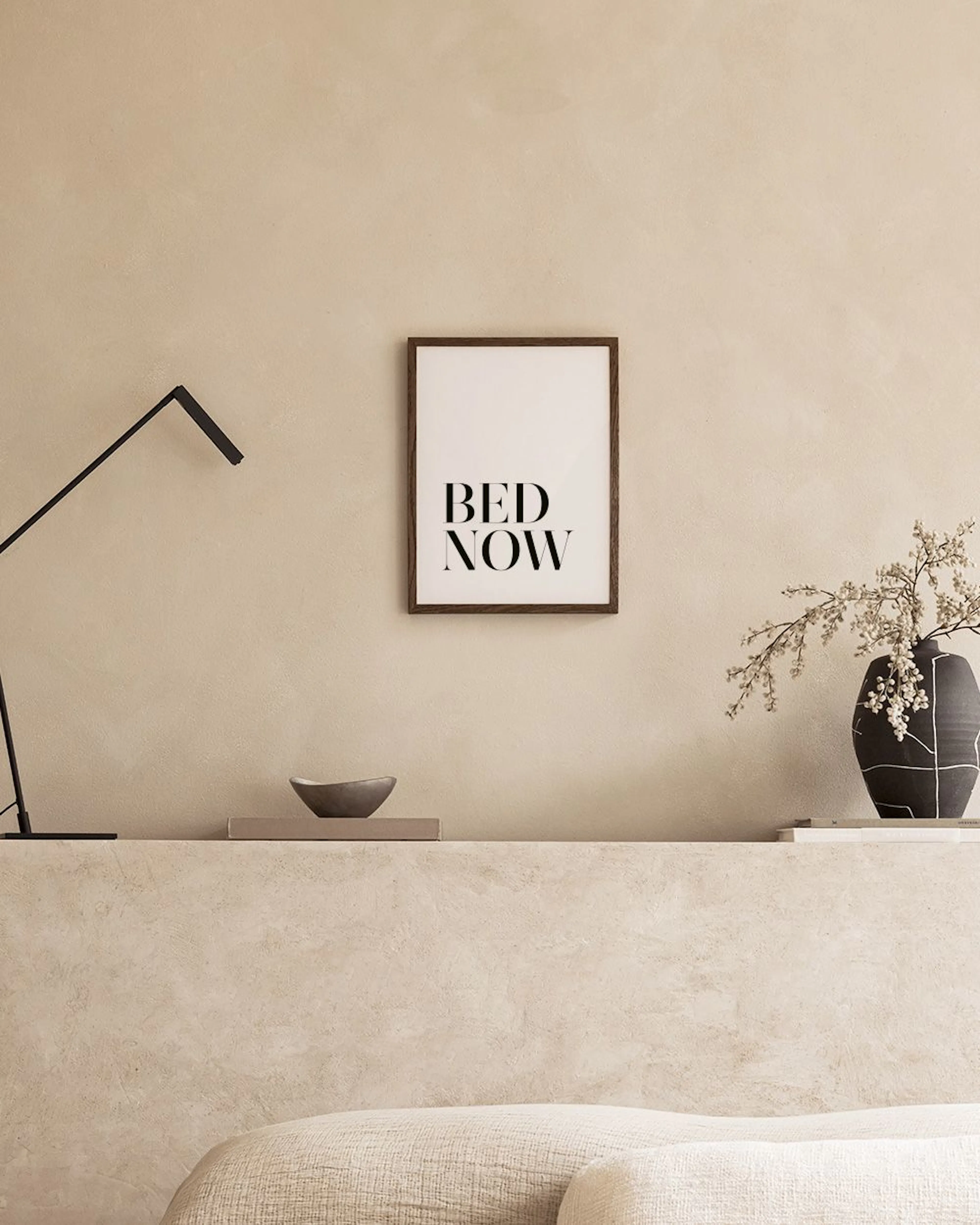 Poster con scritta BED NOW in nero su sfondo bianco, appeso sopra un comodino con un vaso e libri in una camera da letto.
