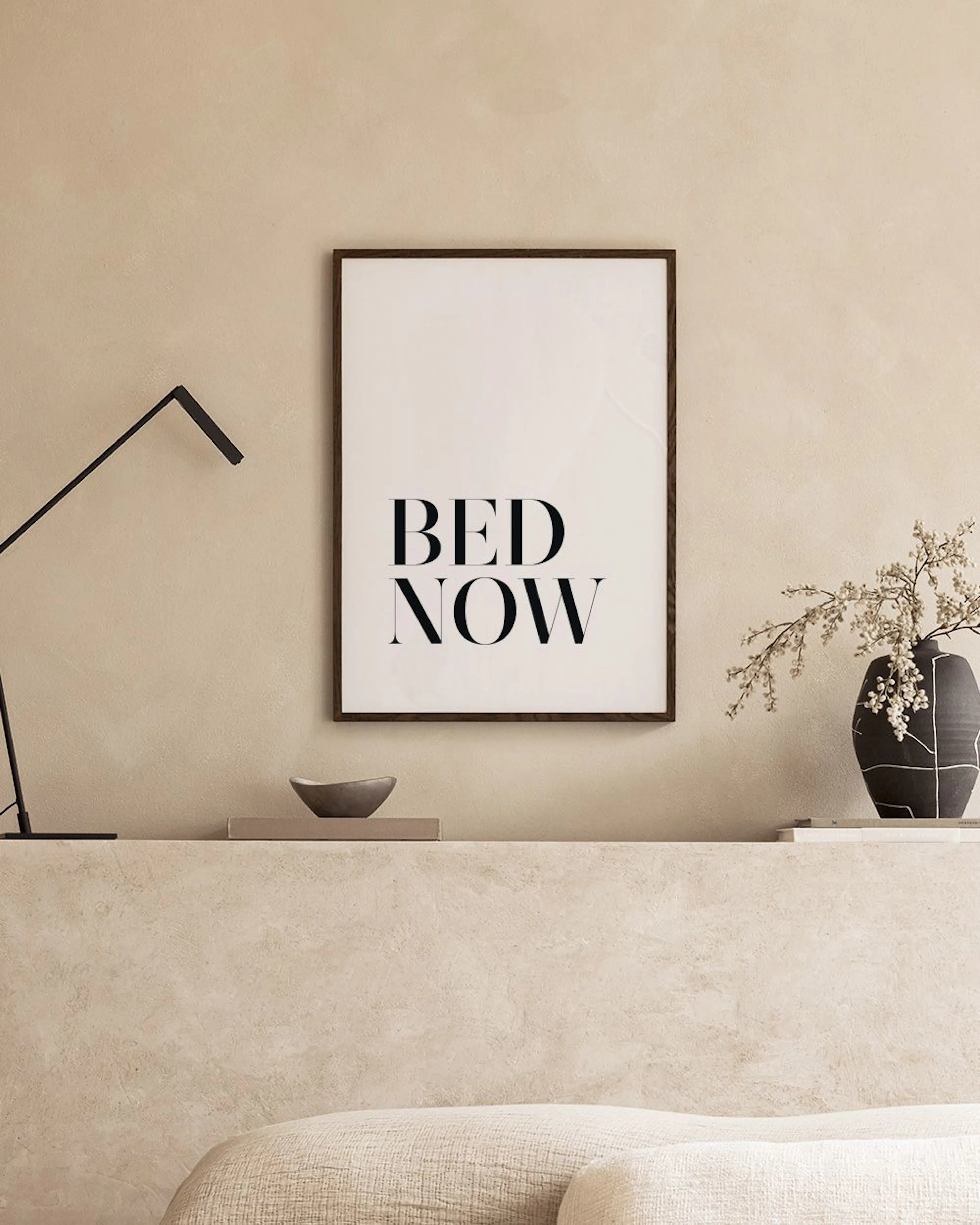 Kamerposter met BED NOW in zwarte letters, hangend boven een wit nachtkastje met vaas en boeken naast een grijs bed.