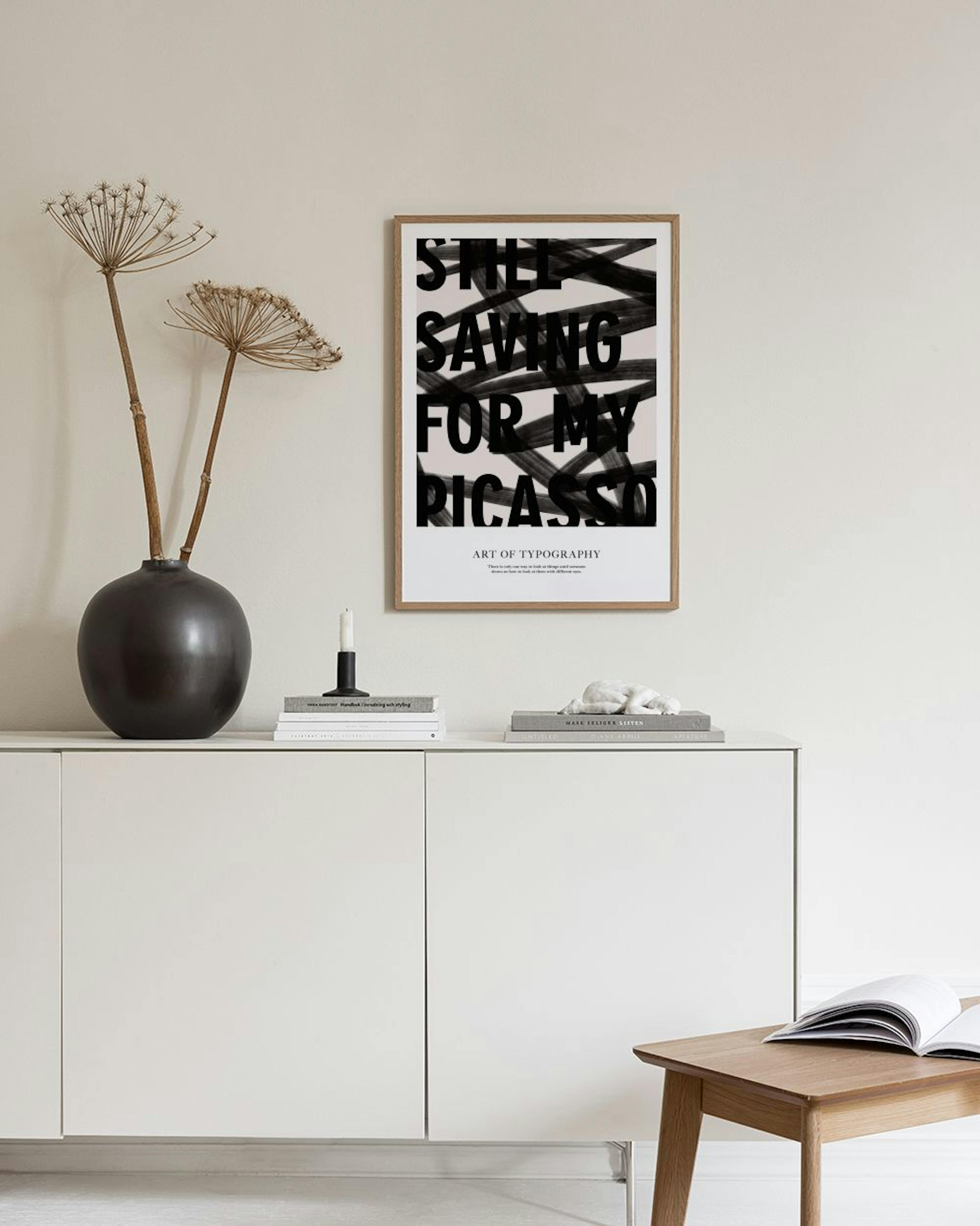Plakat med sort tekst STILL SAVING FOR MY PICASSO over sorte penselstrøg, hængende over et sort bord med en lampe og dekoratione
