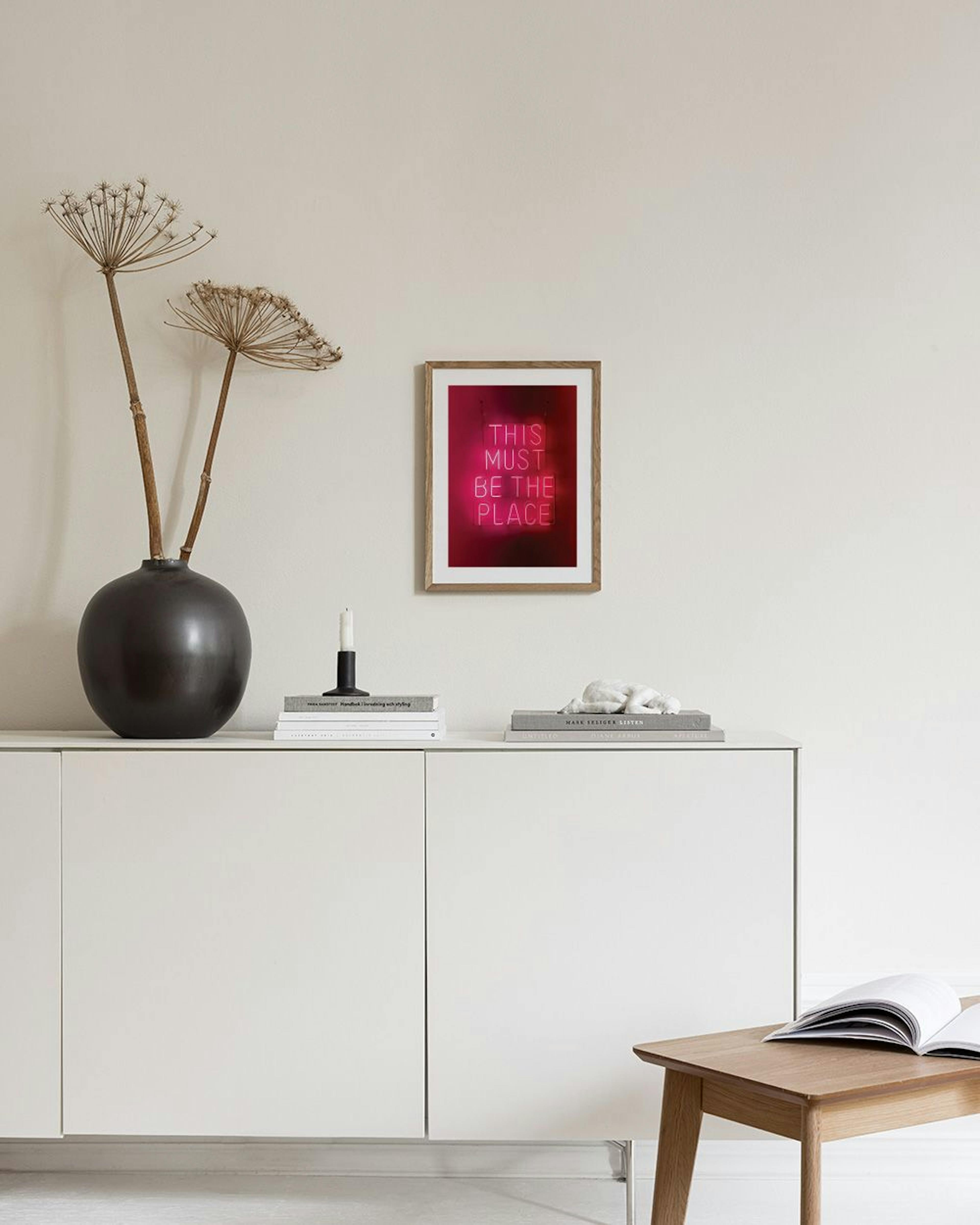 A neon pink print with THIS MUST BE THE PLACE text, hanging above a red tufted ottoman in a modern room.