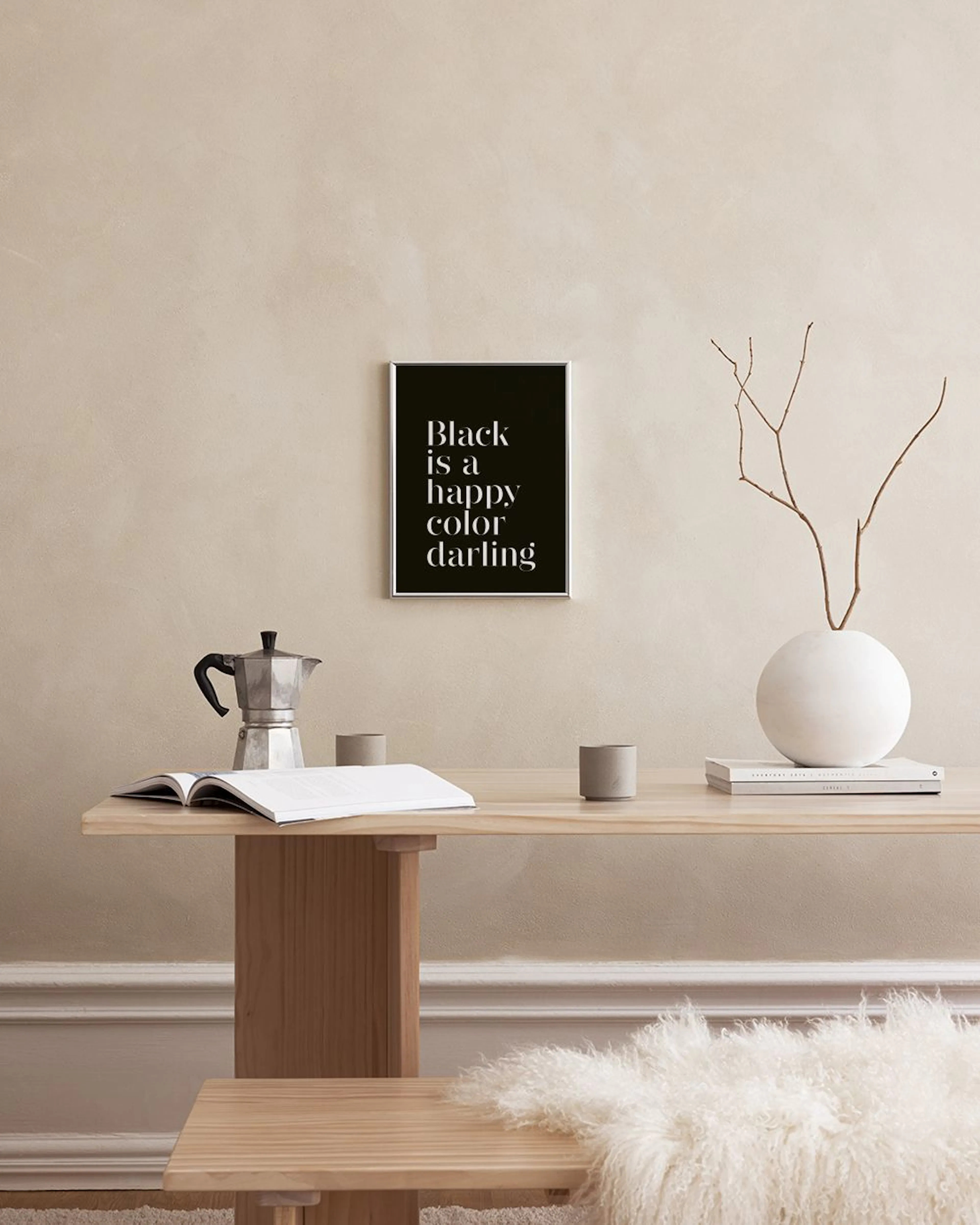 Svart poster med vit text Black is a happy color darling, hängande ovanför ett vitt skåp med böcker, skål och lampa.