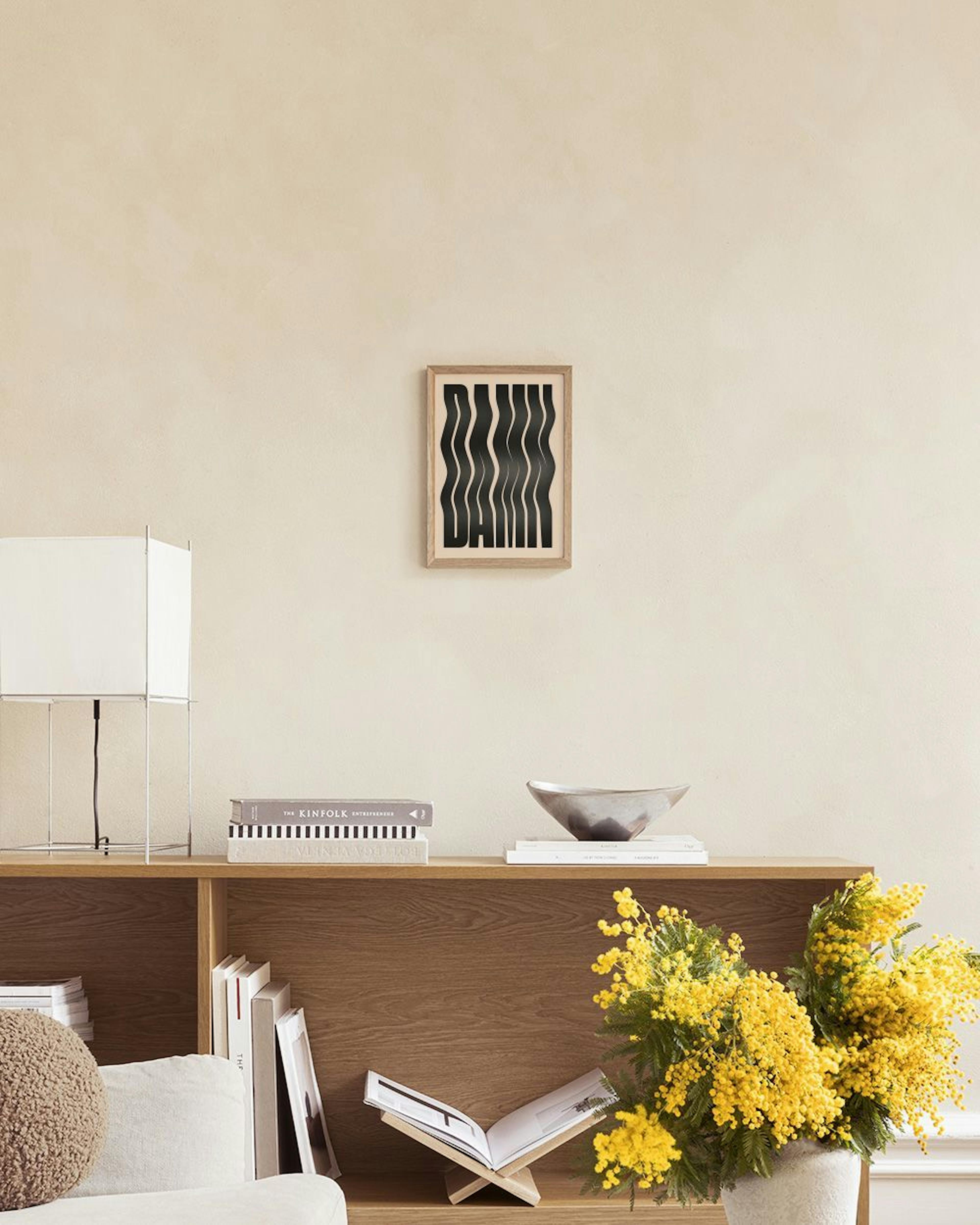 Póster Damn com letras pretas onduladas sobre fundo bege, exibido numa parede cinza clara com uma mesa lateral e vasos.