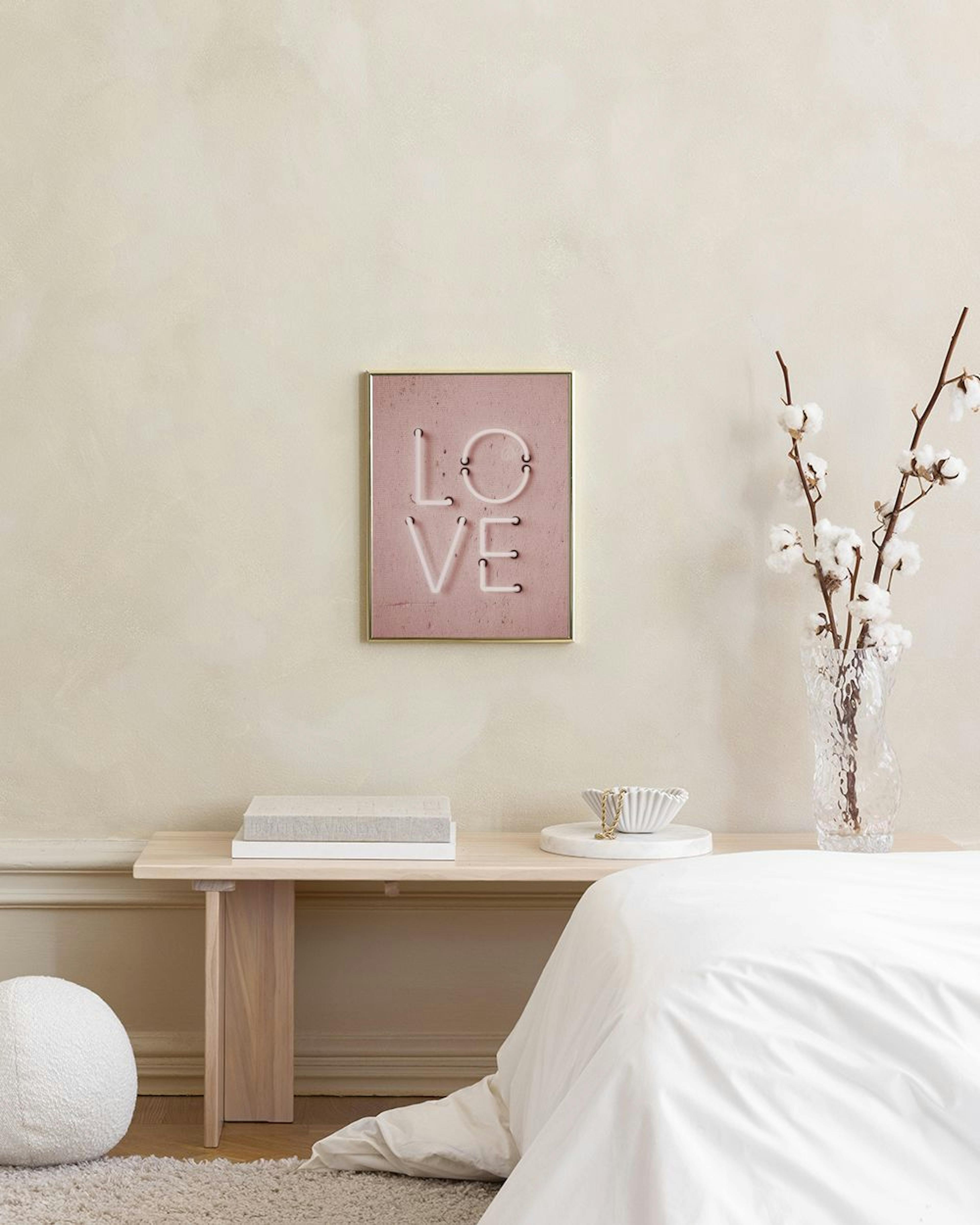 A print of the word LOVE in white neon on a pink concrete background, hanging above a white cabinet with a lamp and books.