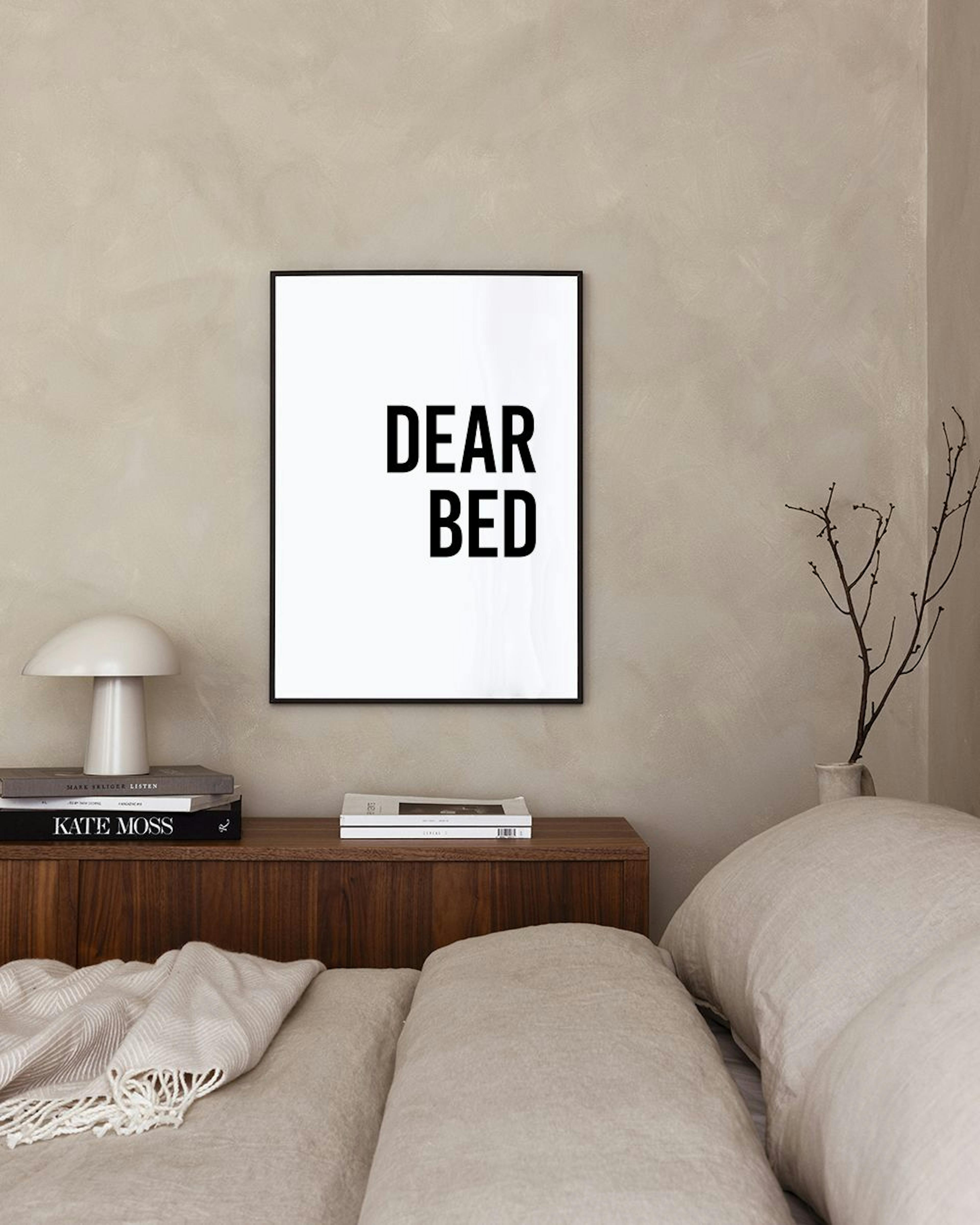A print with DEAR BED in black text on a white background, hanging above a white nightstand in a bedroom.