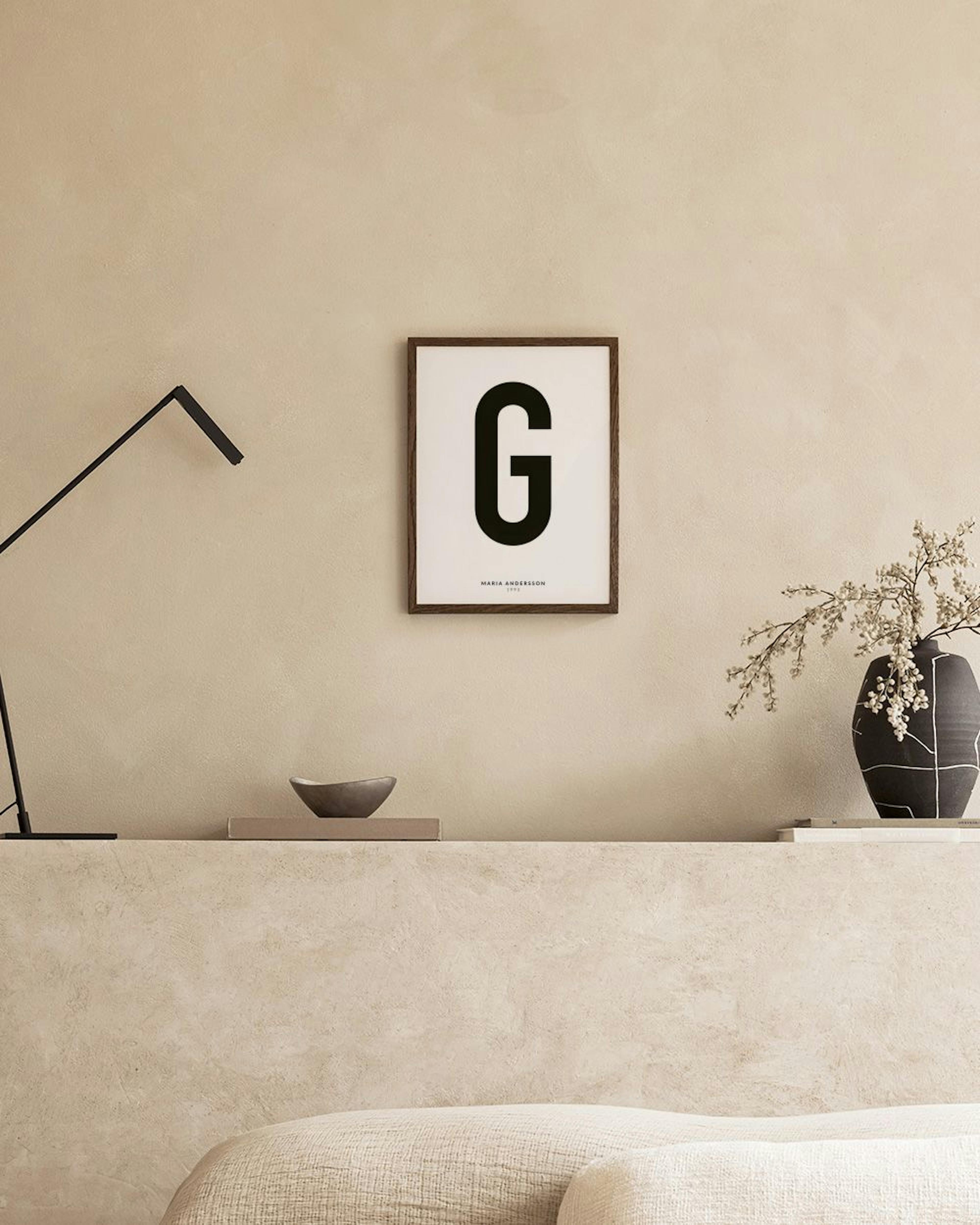 A personalised print with a large black letter G and text MARIA ANDERSSON 1993 hanging above a white cabinet with decor.