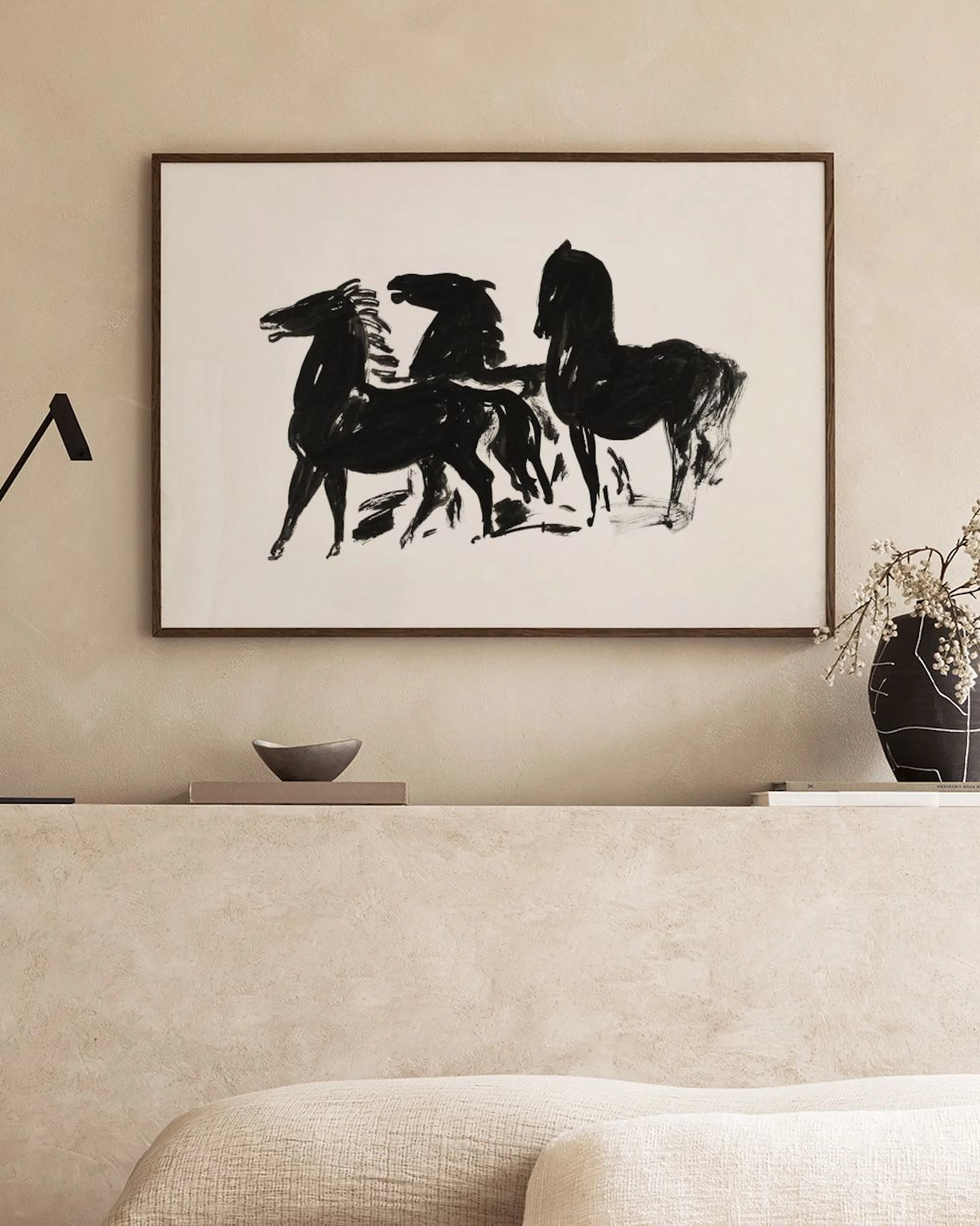 A print of three black horses in an expressive, painterly style, hanging above a bed with white and beige pillows.
