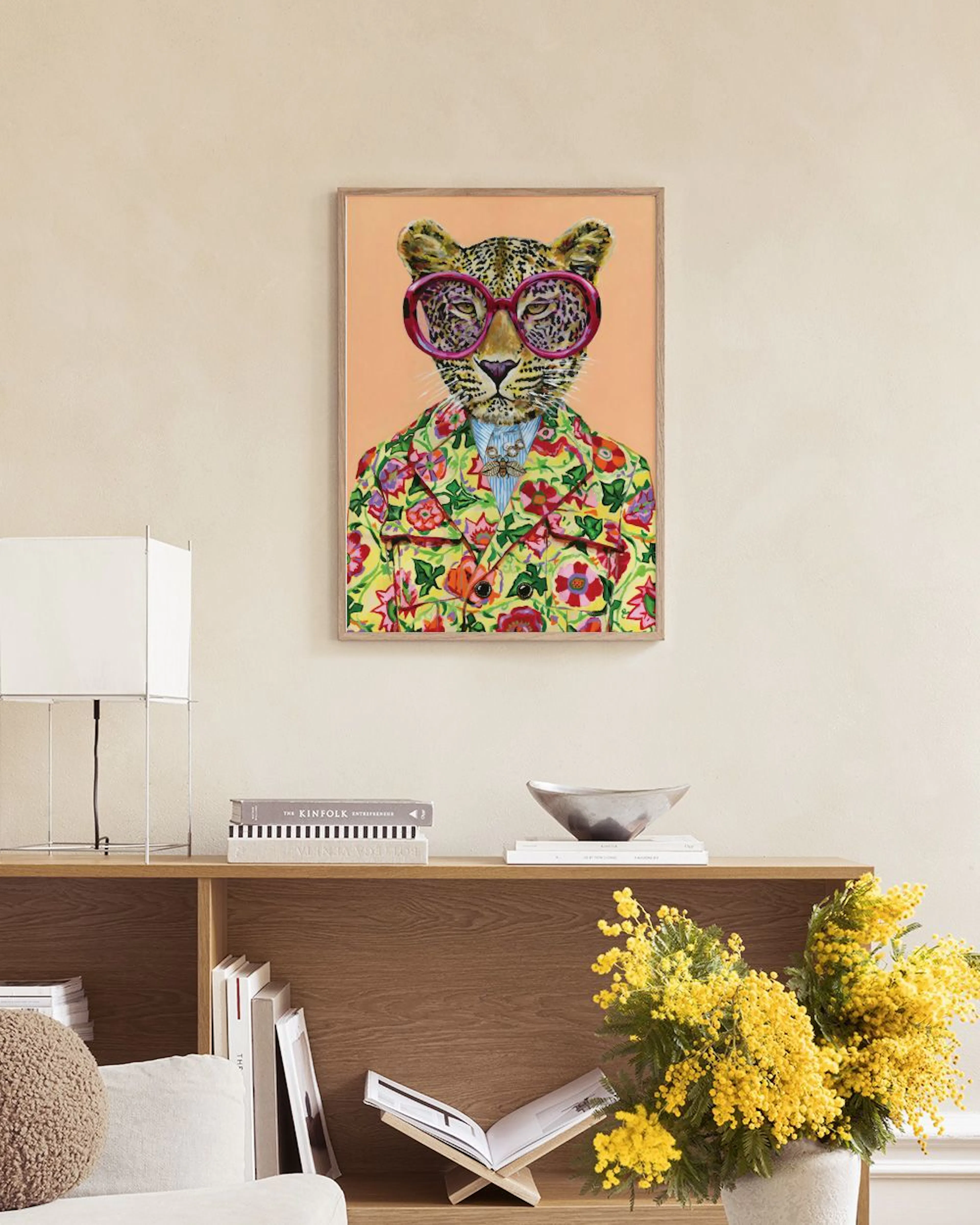 Poster eines Leoparden mit pinker Brille und bunter Blumenjacke, hängt über einem pinken Sofa mit Kissen und Decke.