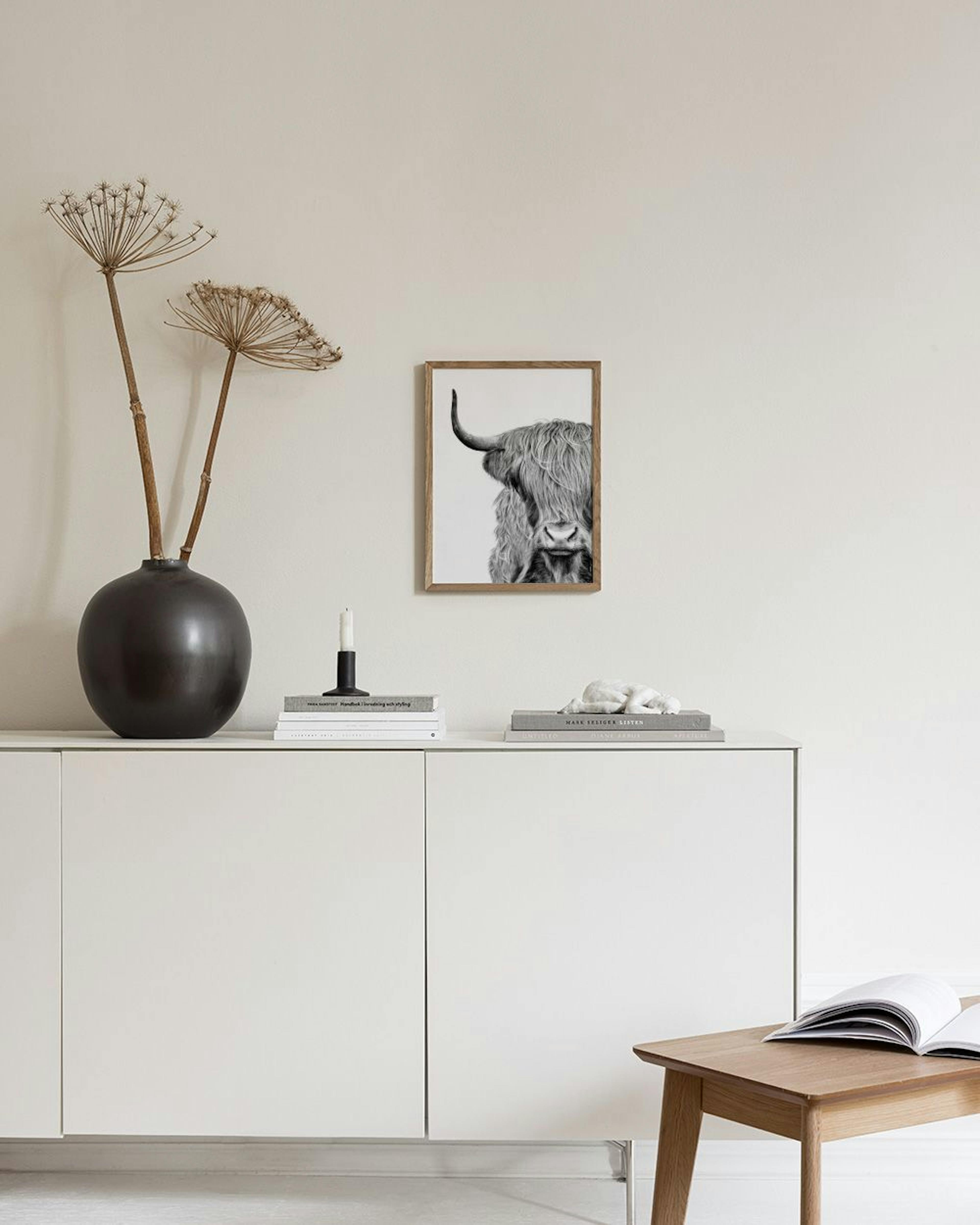 A black and white print of a Highland cow’s head, with shaggy hair covering its eyes, hanging above a small stool with books.
