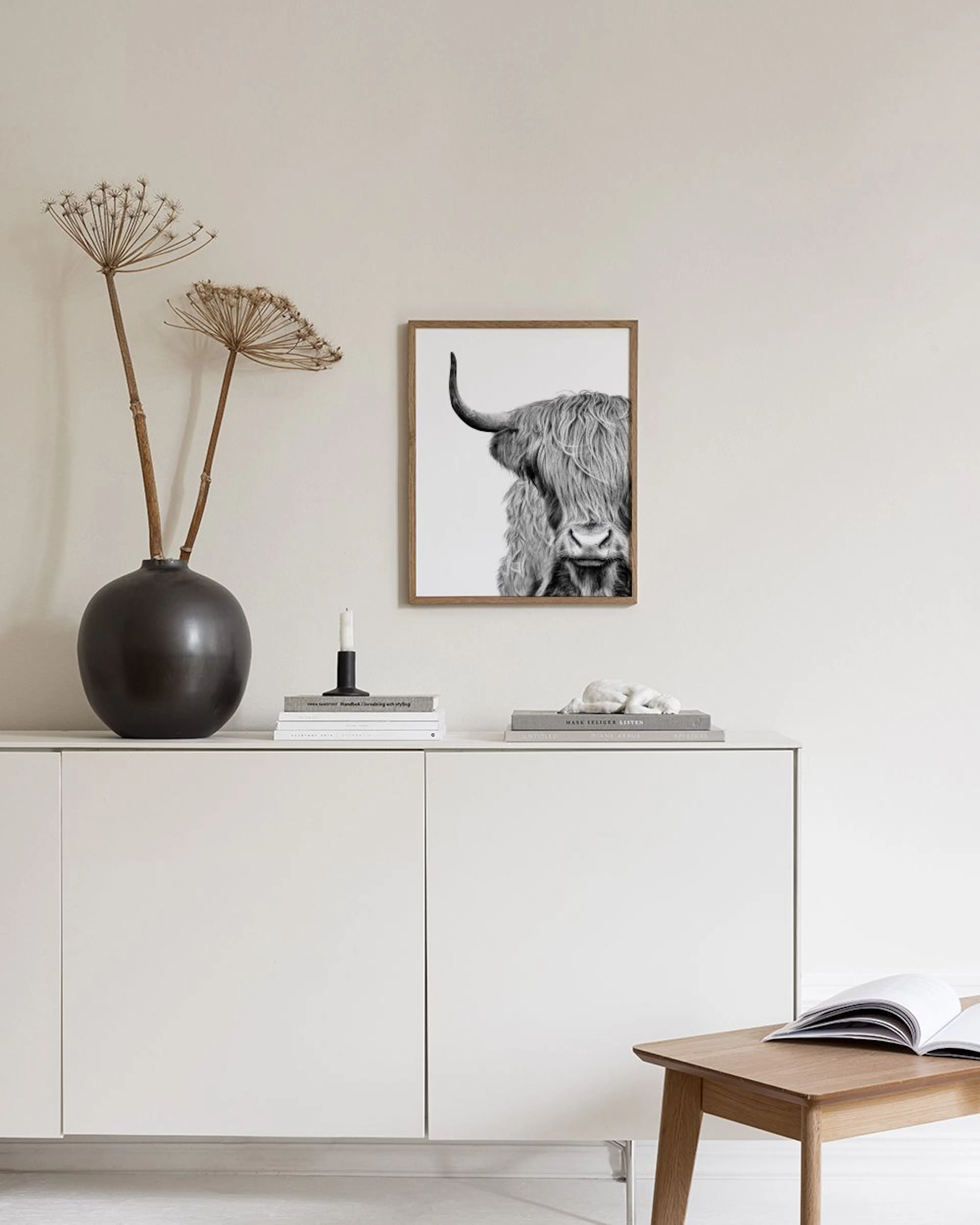 Schwarz-weiß Poster einer Highland-Kuh, die an einer weißen Wand über einem Hocker mit Büchern hängt.