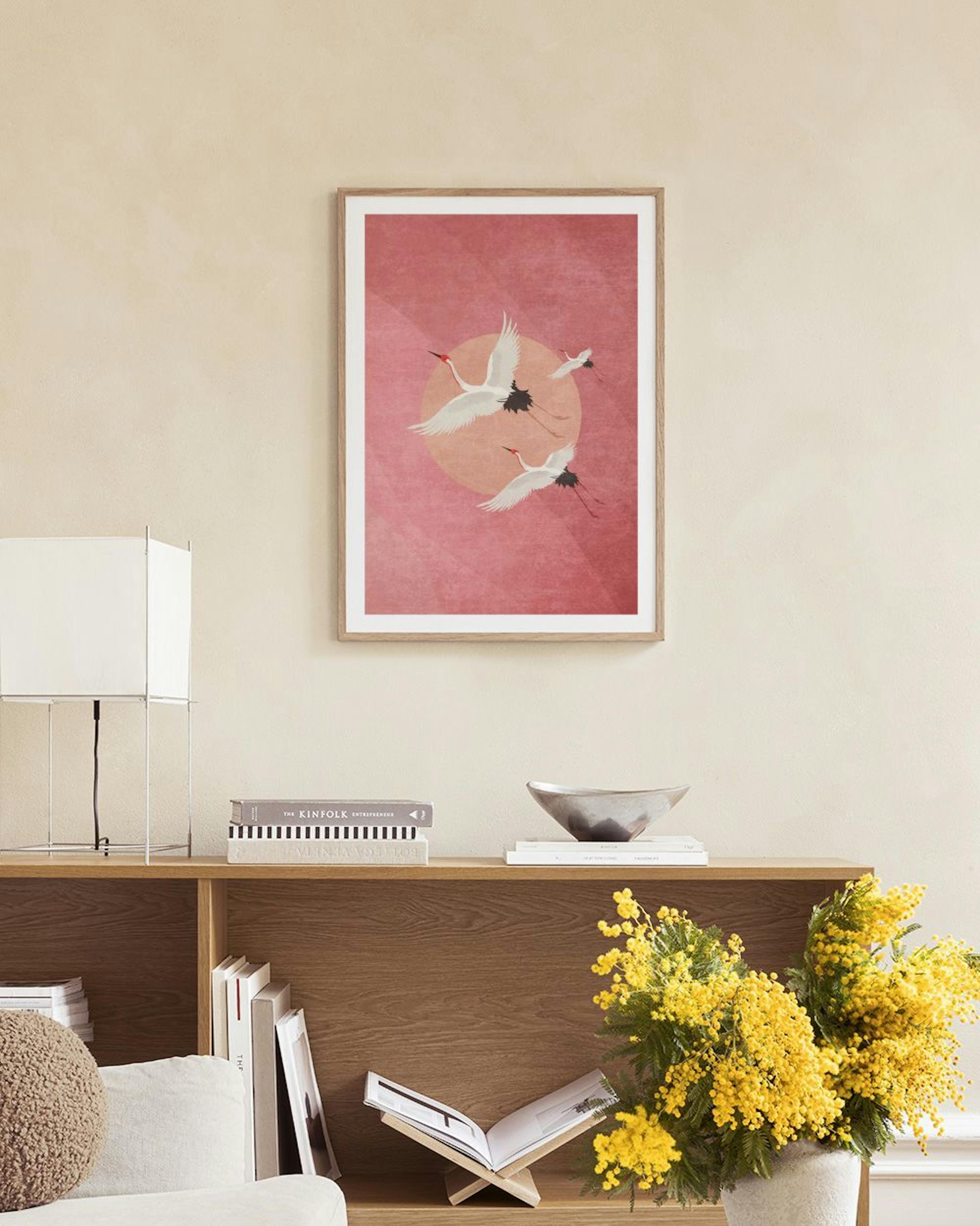 Poster met drie vliegende kraanvogels voor een oranje zon op een roze achtergrond, staand op een grijze bank met decoratie en la