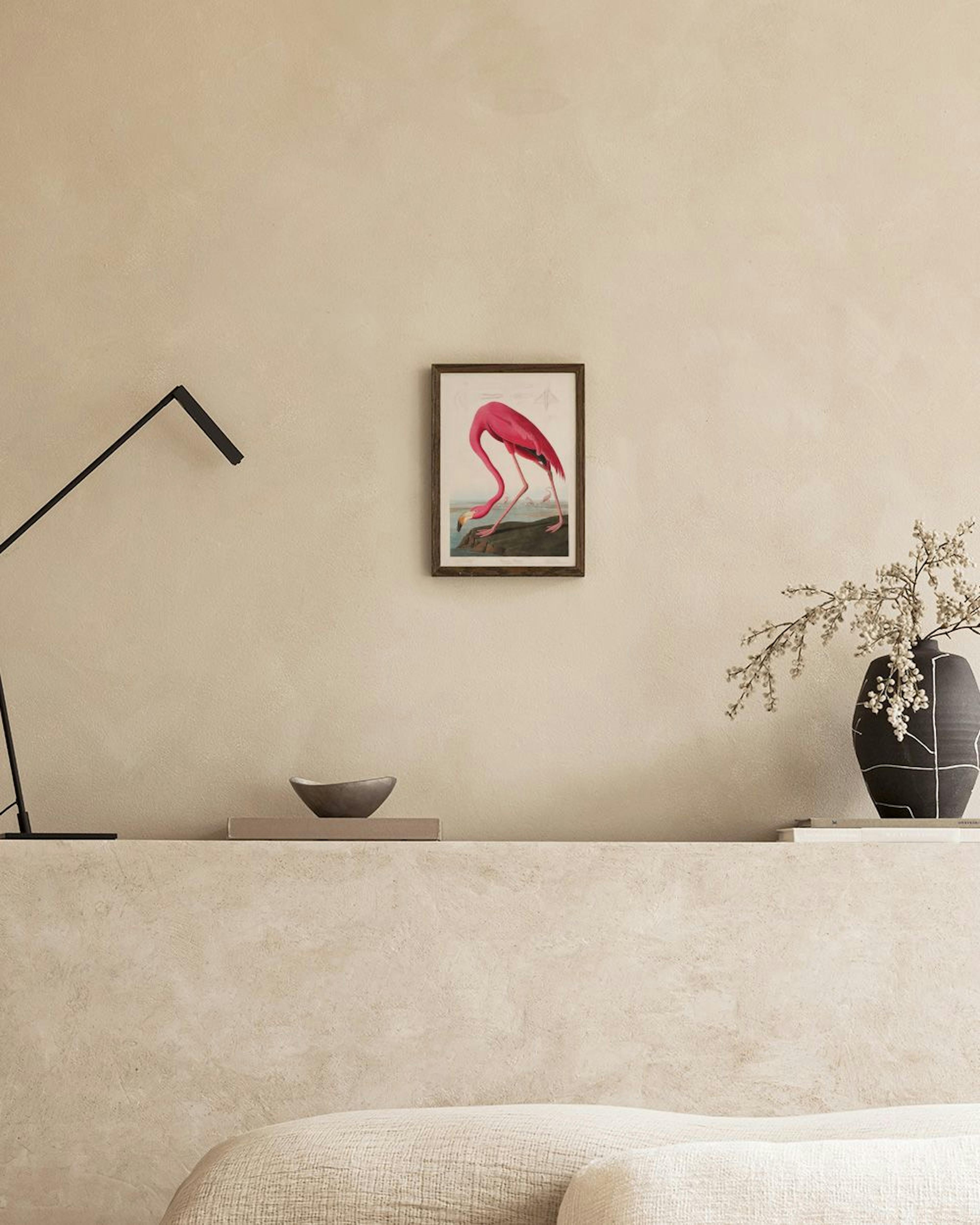 A vibrant pink flamingo print, with long legs and a curved neck, bending to drink from water, displayed above a black console ta