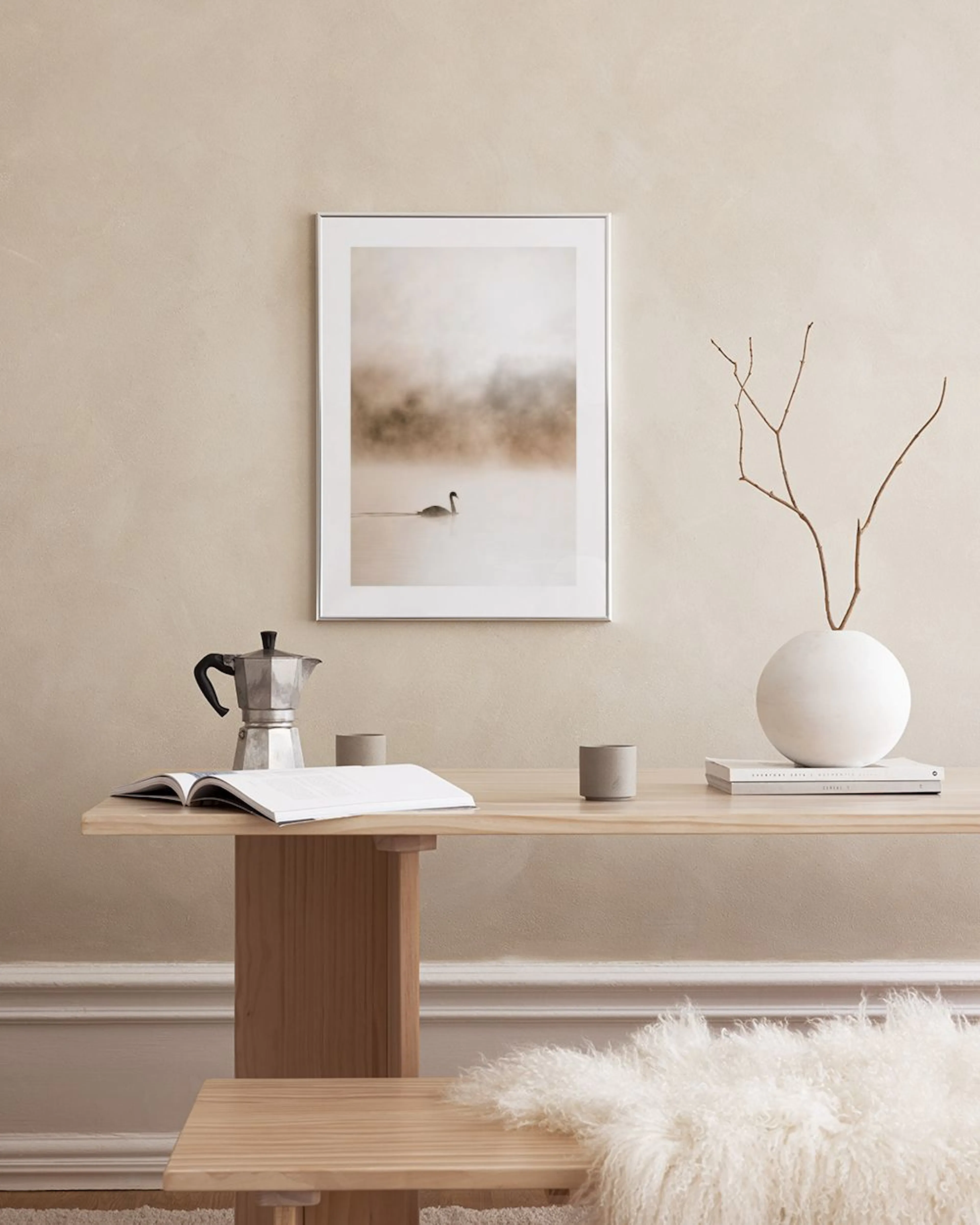 A print of a swan swimming in a misty, light brown and white lake, hanging on a beige wall above a black table and brown ottoman