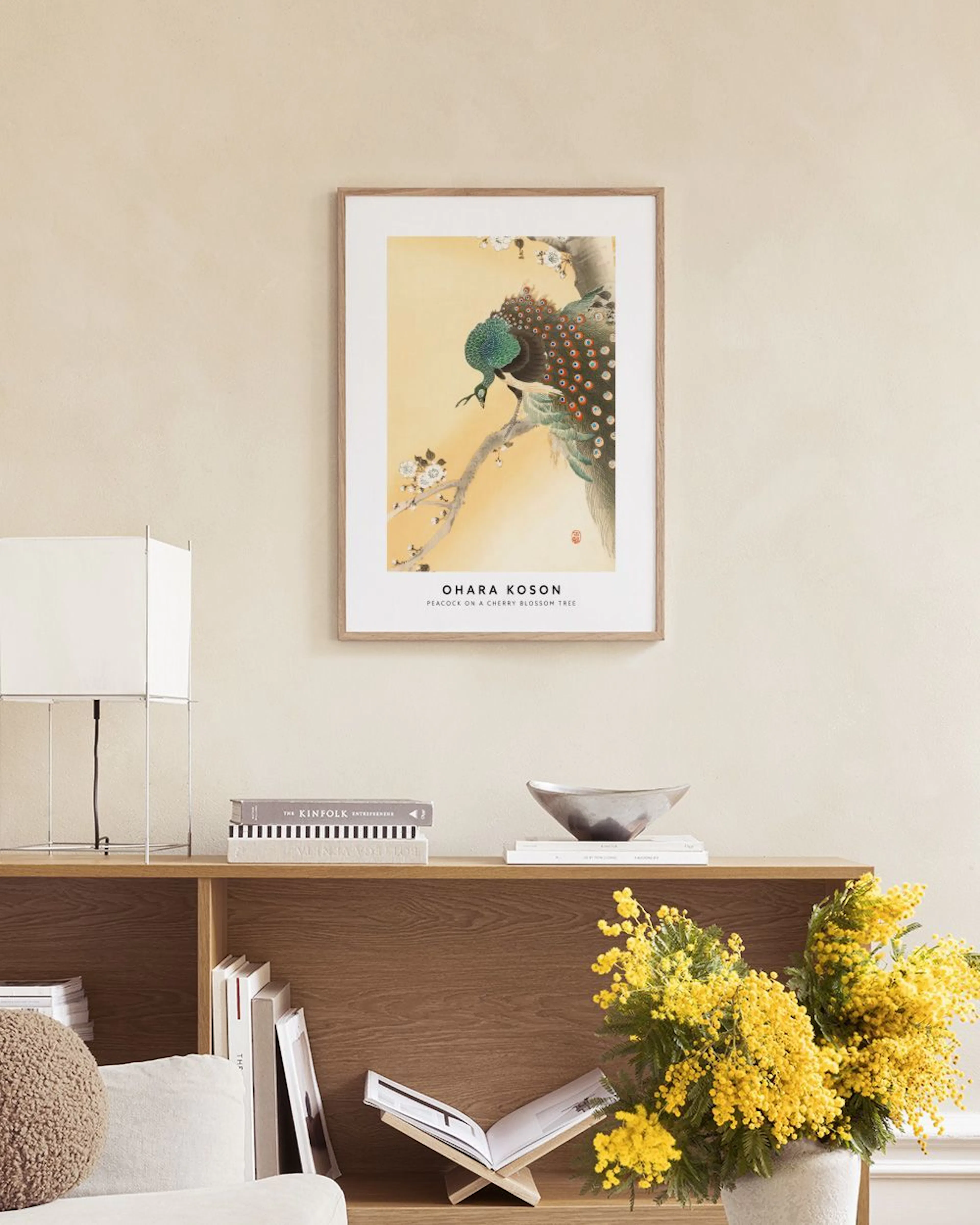 A peacock print with a cherry blossom branch on a yellow background, hanging above a side table in a cozy living room.