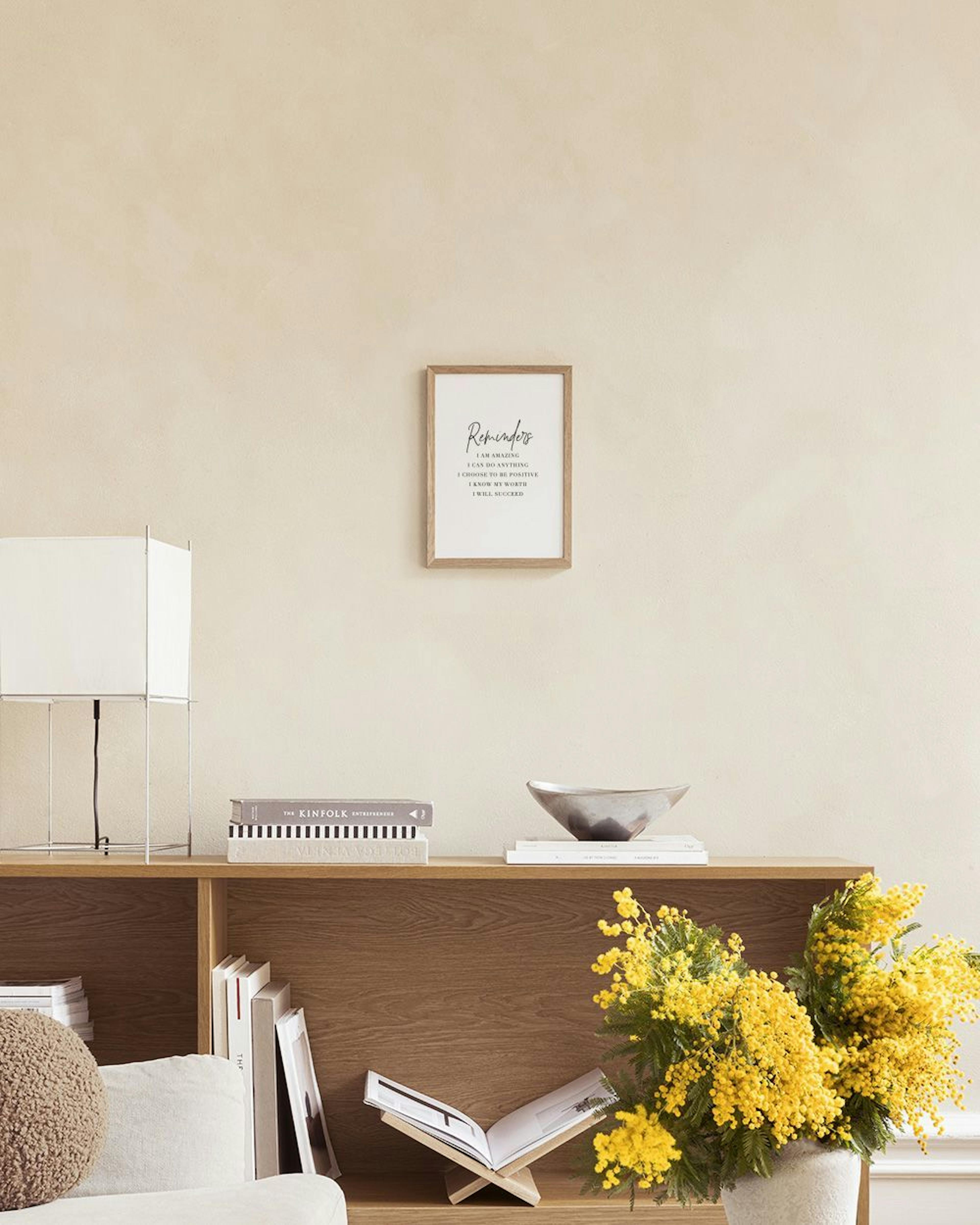 Póster de texto con Reminders y afirmaciones en español, sobre una cómoda beige con jarrón y planta.