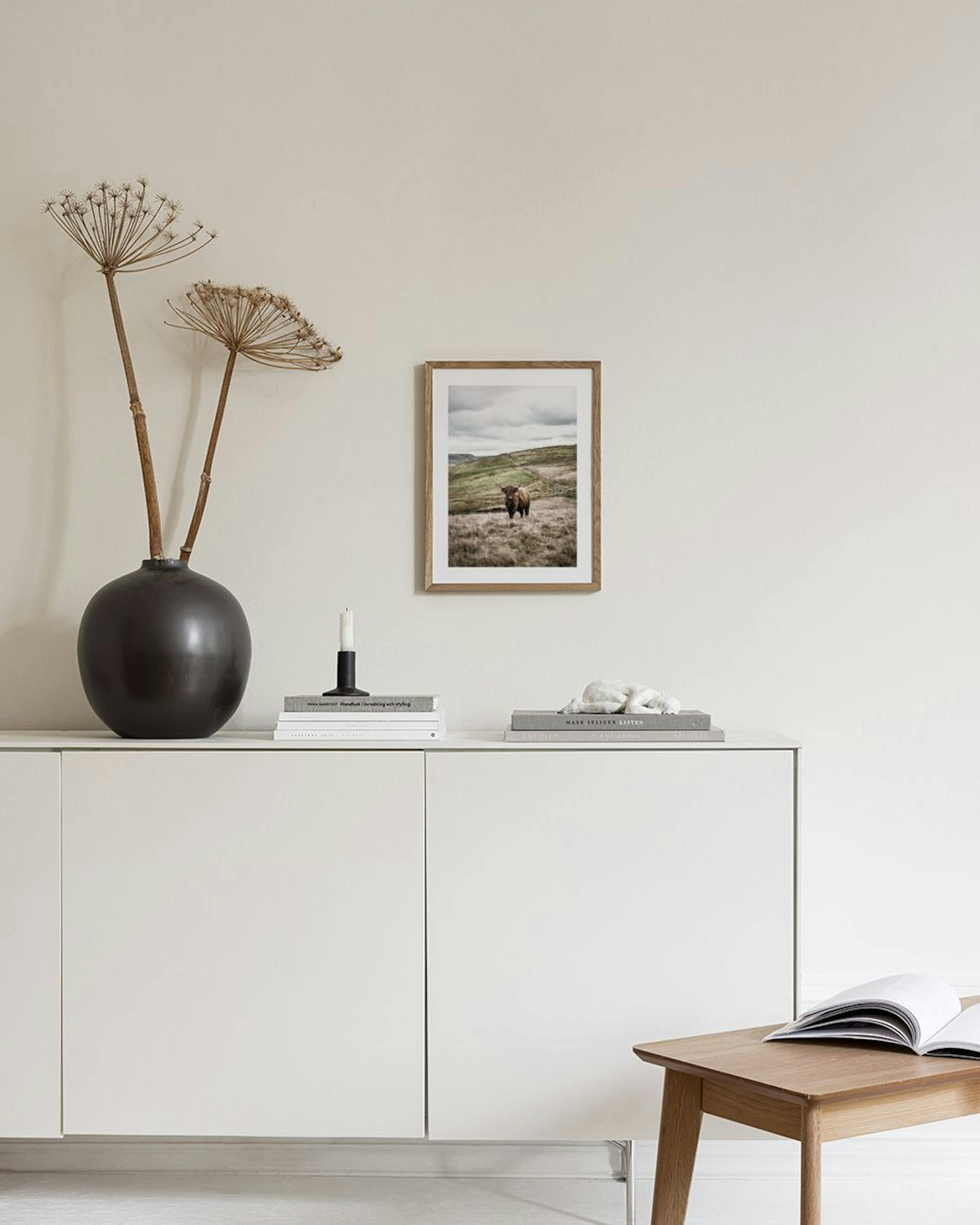 Poster einer Schottischen Hochlandkuh auf einer Wiese, gerahmt an einer grauen Wand über einem weißen Sideboard mit Vase.