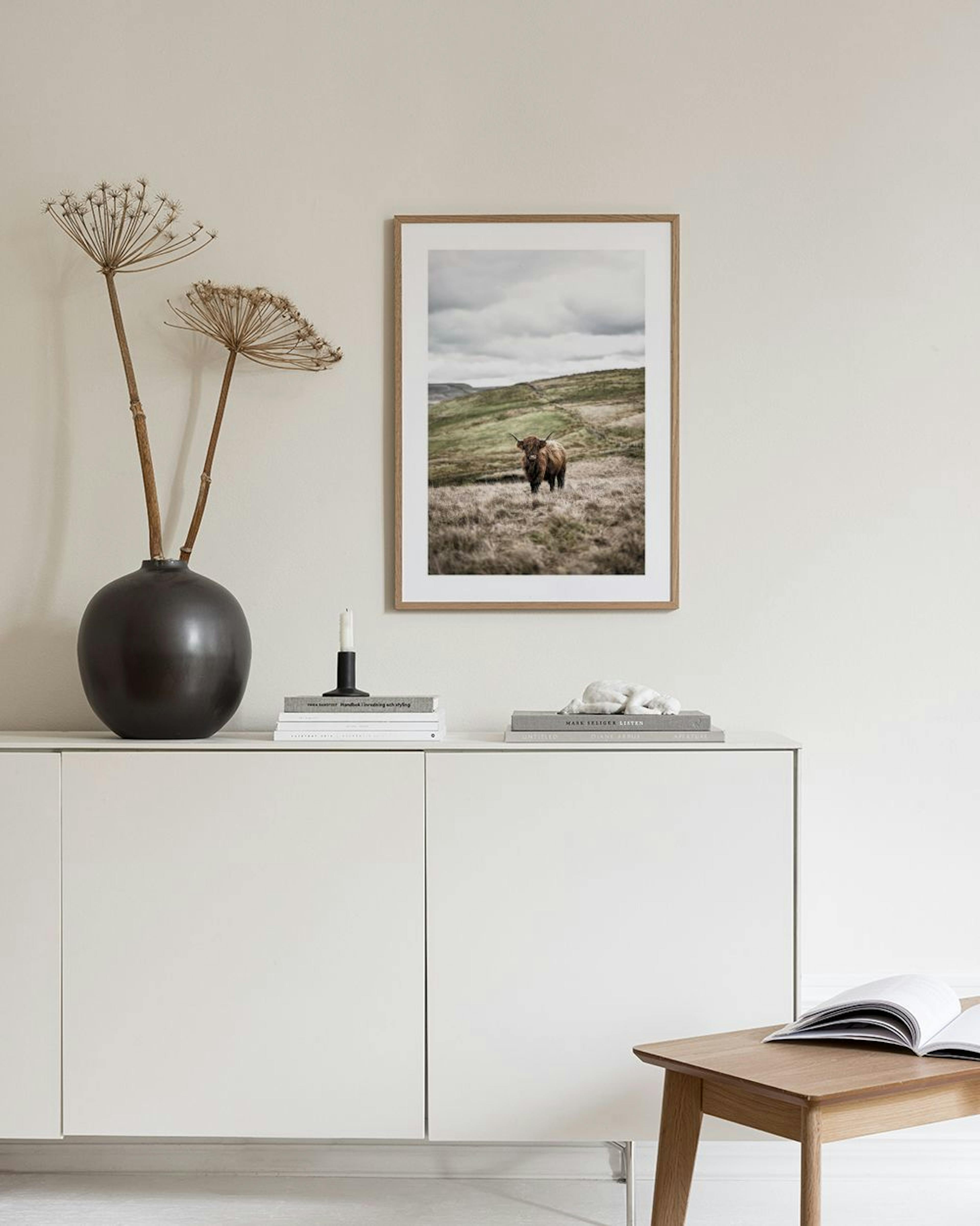 A poster of a brown Highland cow in a grassy field with hills under a cloudy sky, hanging on a gray wall above a white cabinet.