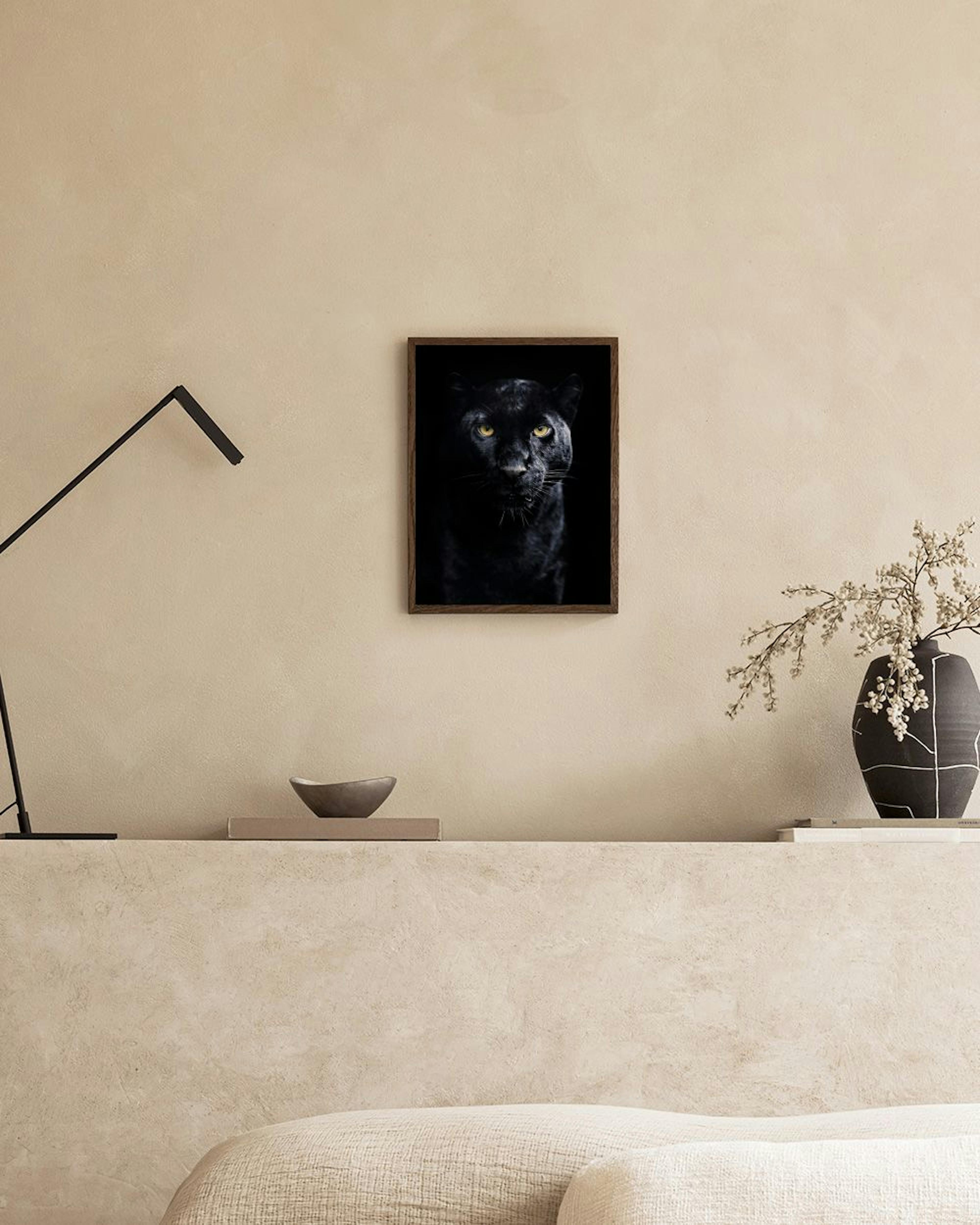 A black panther print with piercing yellow eyes, hanging on a dark wall next to a gold lamp and blue velvet ottoman.