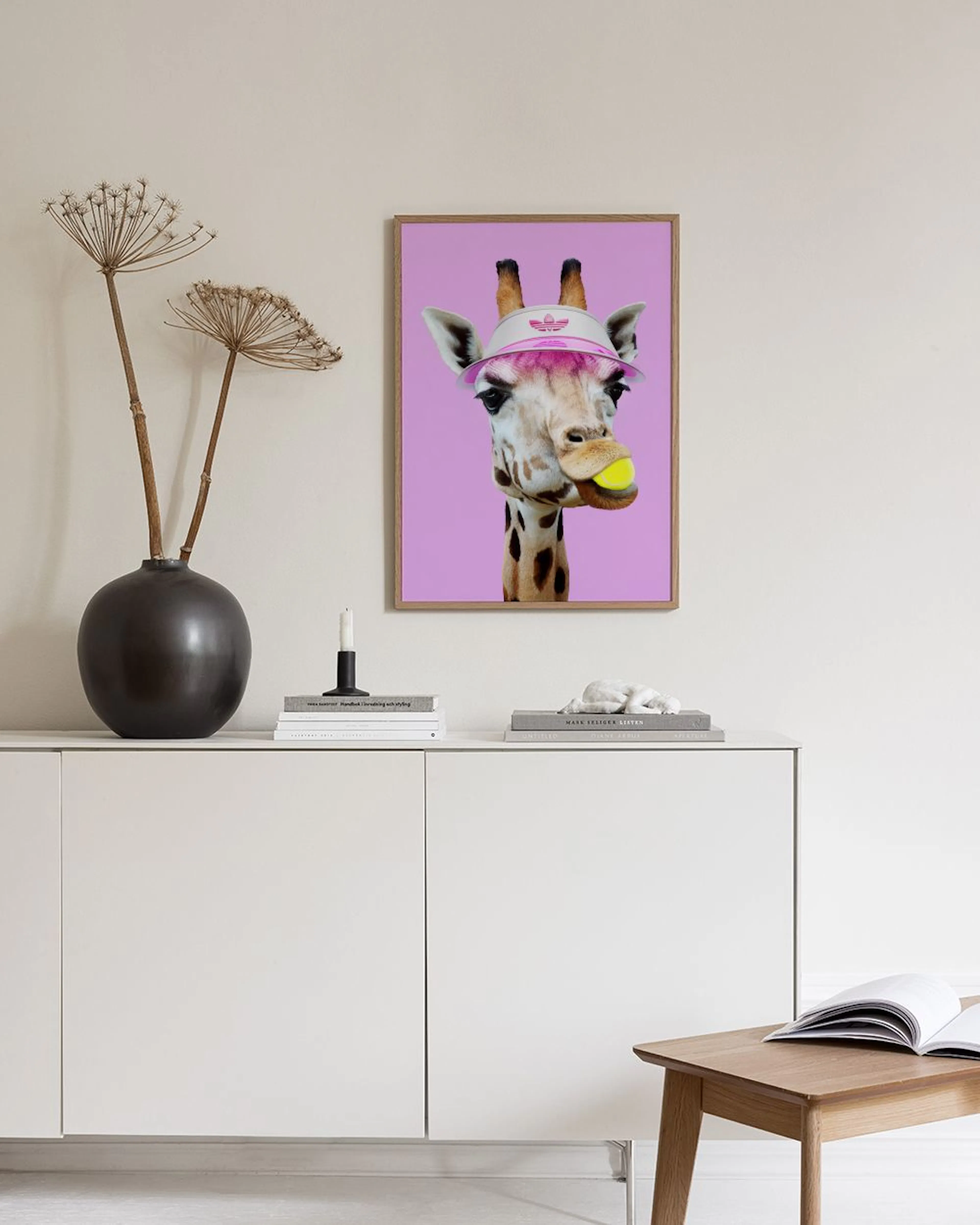 Affiche dune girafe avec une visière rose et une balle de tennis en bouche, accrochée sur un mur blanc au-dessus dun tabouret en