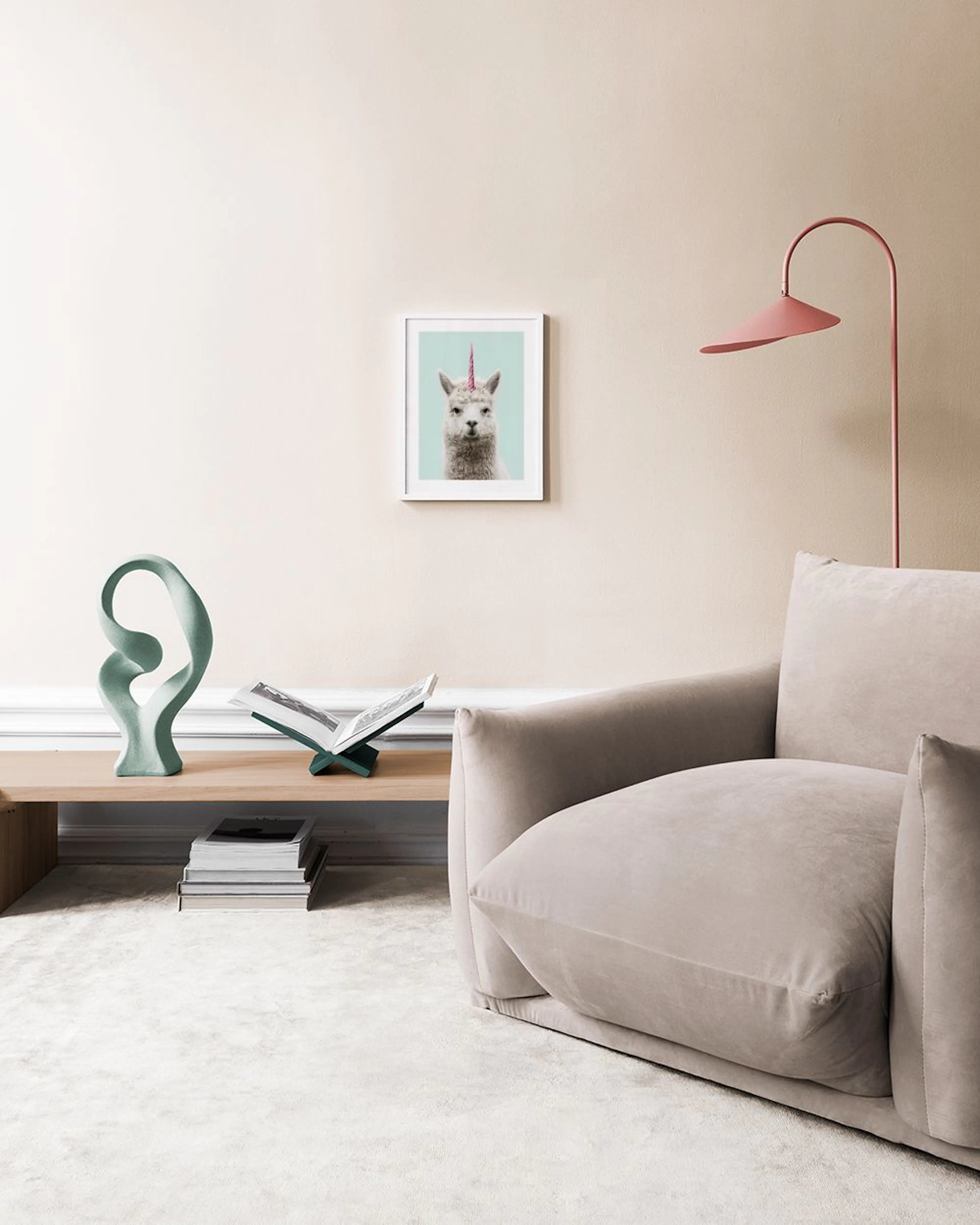 Póster de una alpaca blanca con cuerno rosa de unicornio sobre fondo verde menta, colgado sobre un banco de madera y mimbre.
