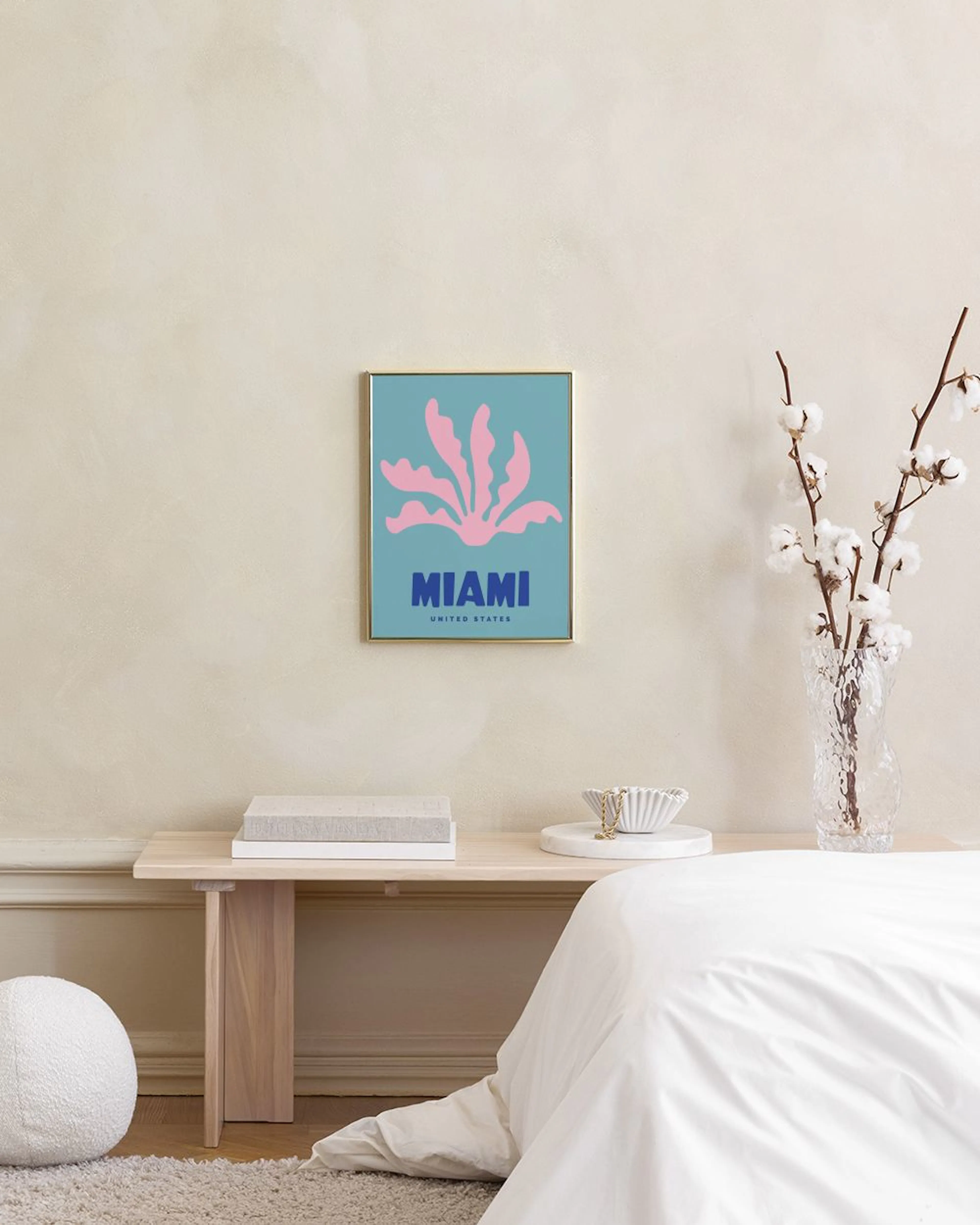 Miami print with pink organic shapes and blue text, hanging above a wooden desk with a cactus and lamp.