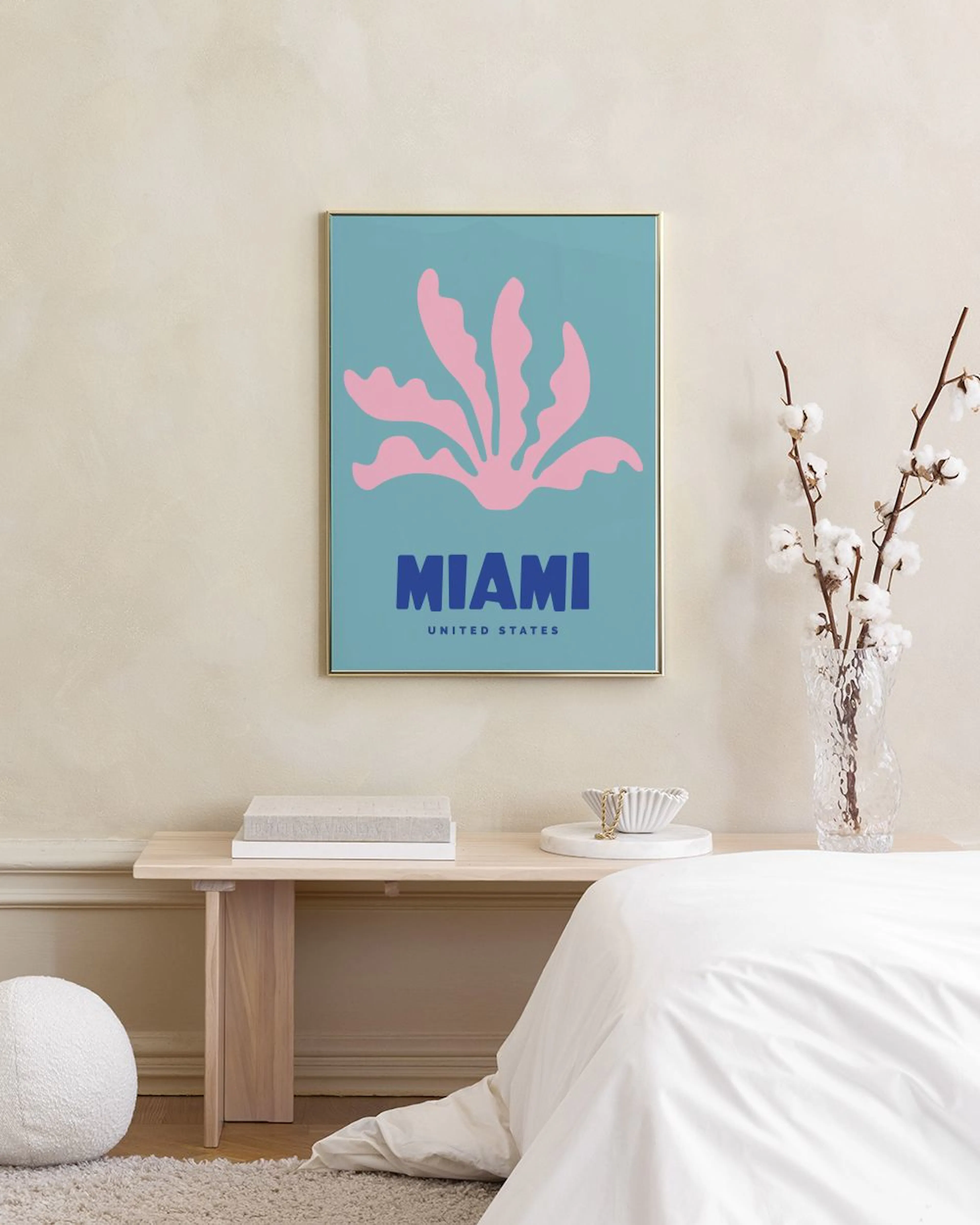 Póster de Miami con formas rosadas y texto azul sobre fondo celeste, colgado sobre un escritorio de madera con cactus y lámpara.