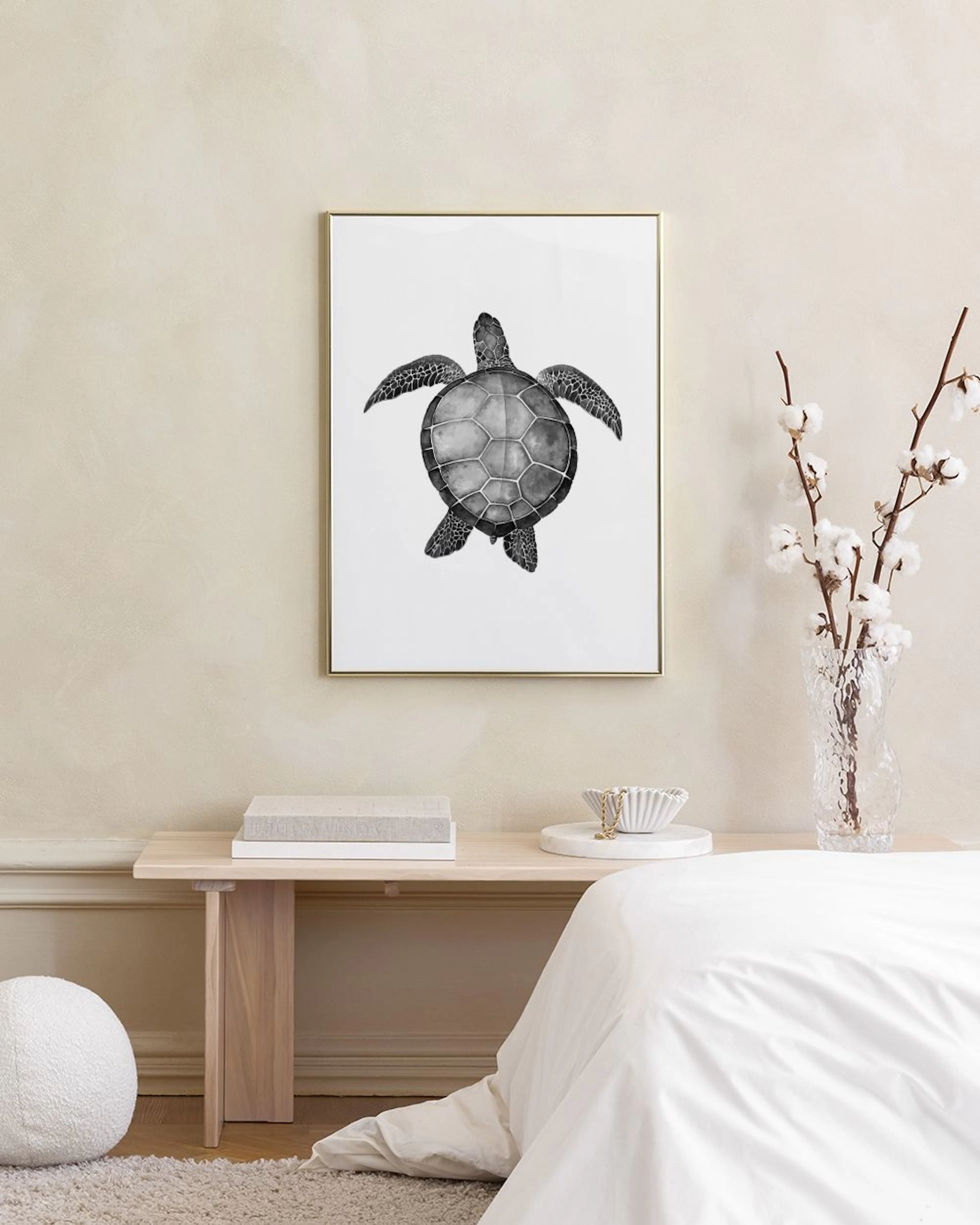 A black and white print of a sea turtle hanging above a side table with books and pink glass vases.