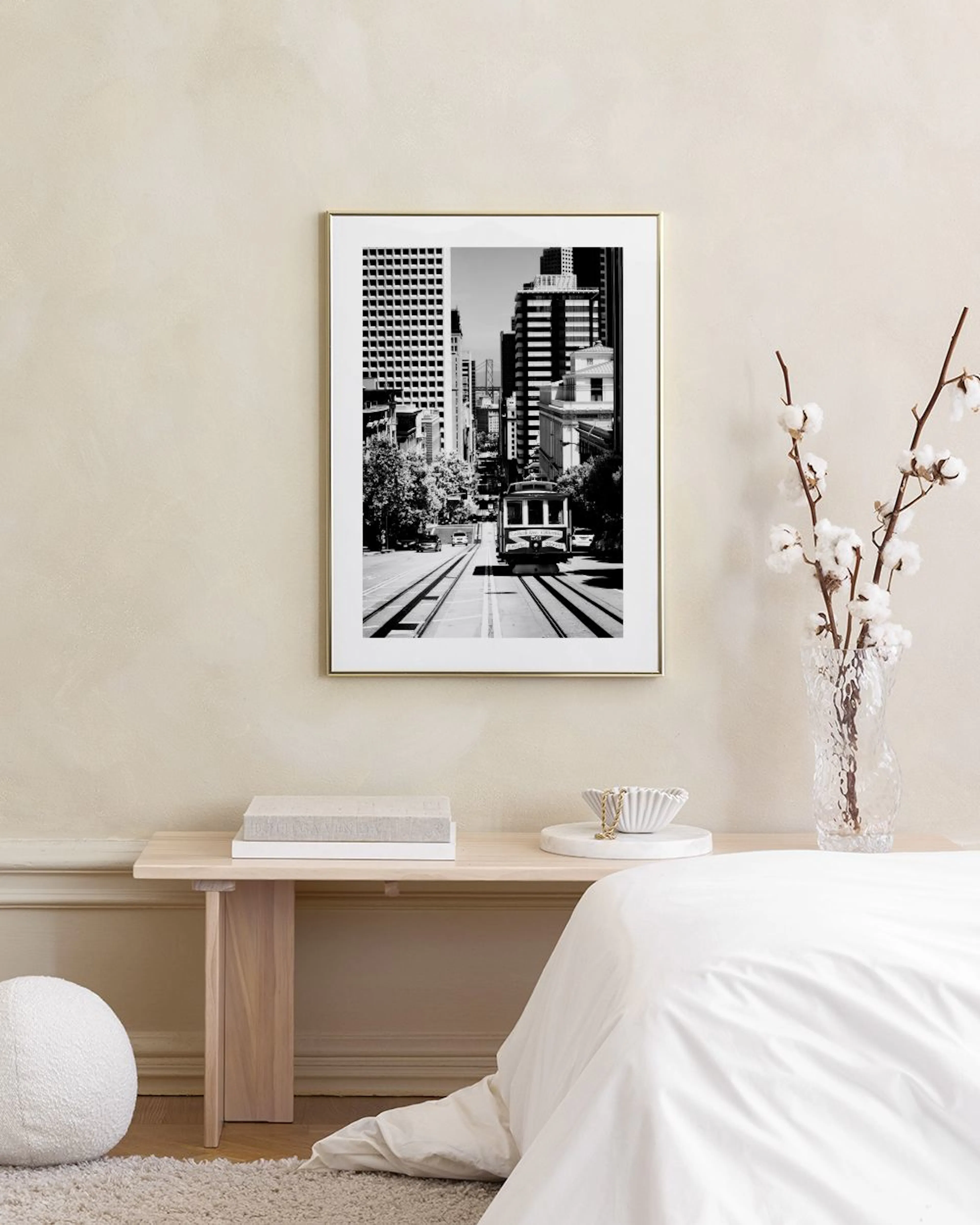 Monochrome print of a city street with a tram and tall buildings, hanging on a white wall above a stool with books.
