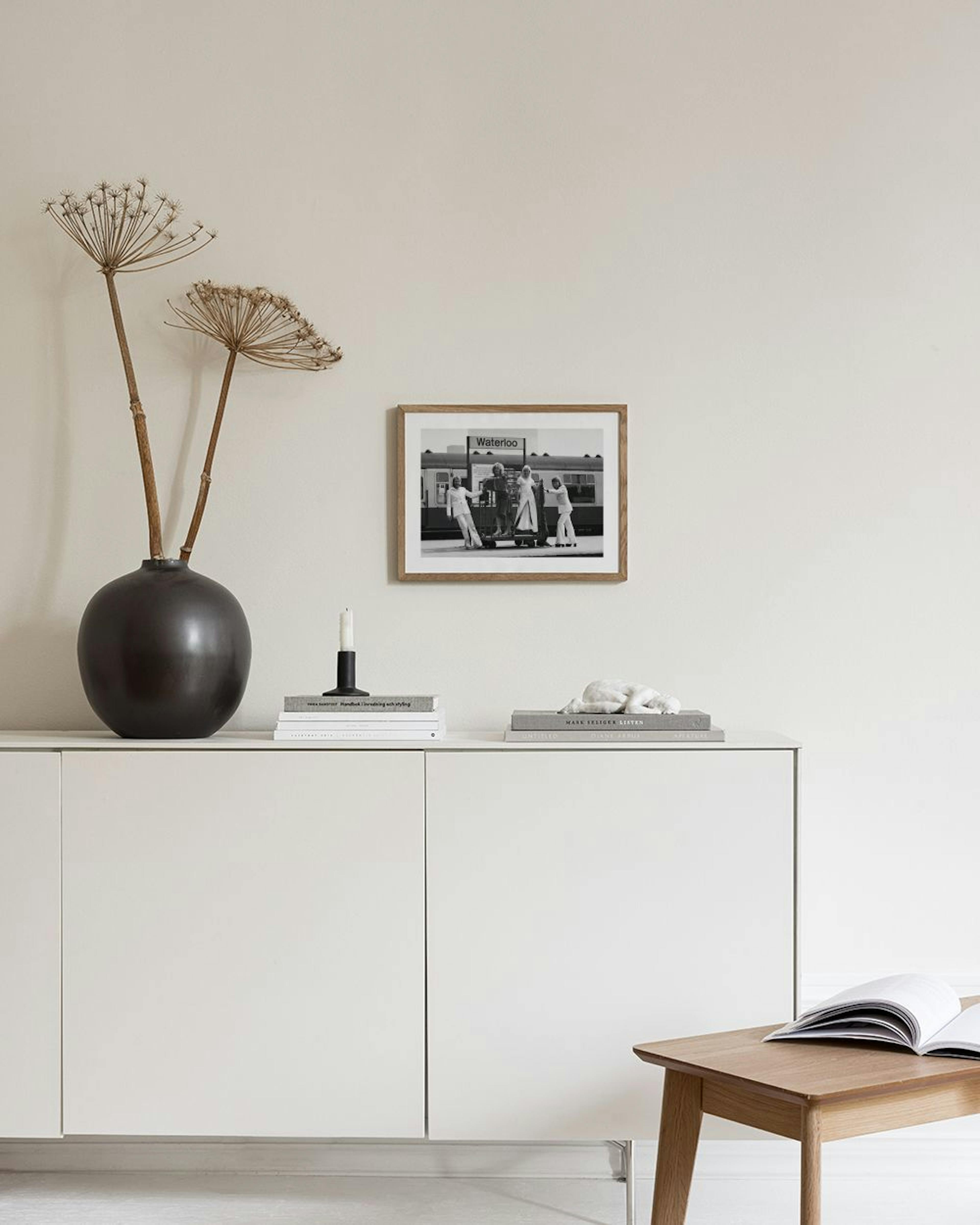 A black and white print of ABBA at the Waterloo train station, displayed above a dark wooden cabinet with vases and books.