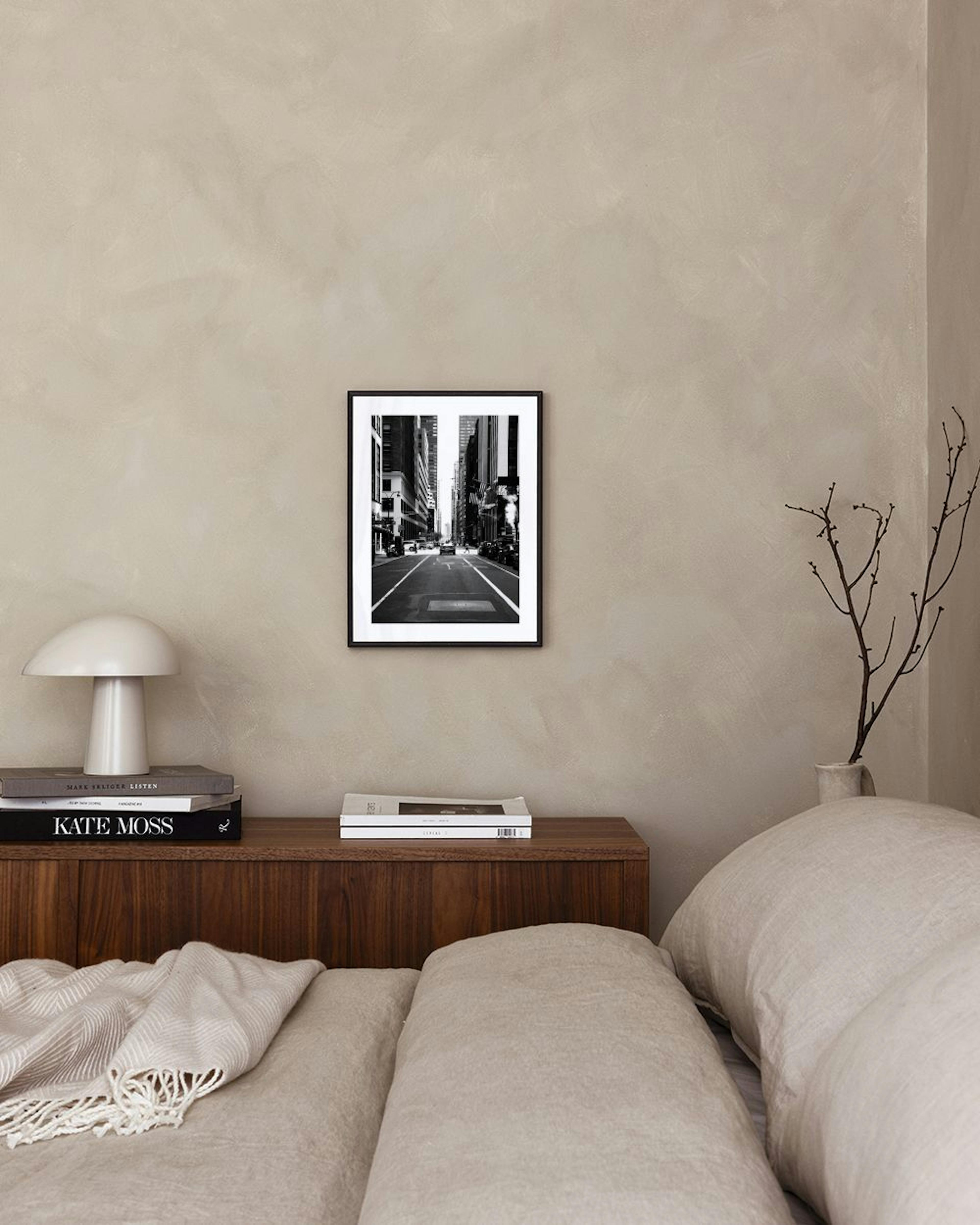 Plakat med sort-hvidt fotografi af en bygade med biler og skyskrabere, hængende over et sort skab med bøger og en lampe.