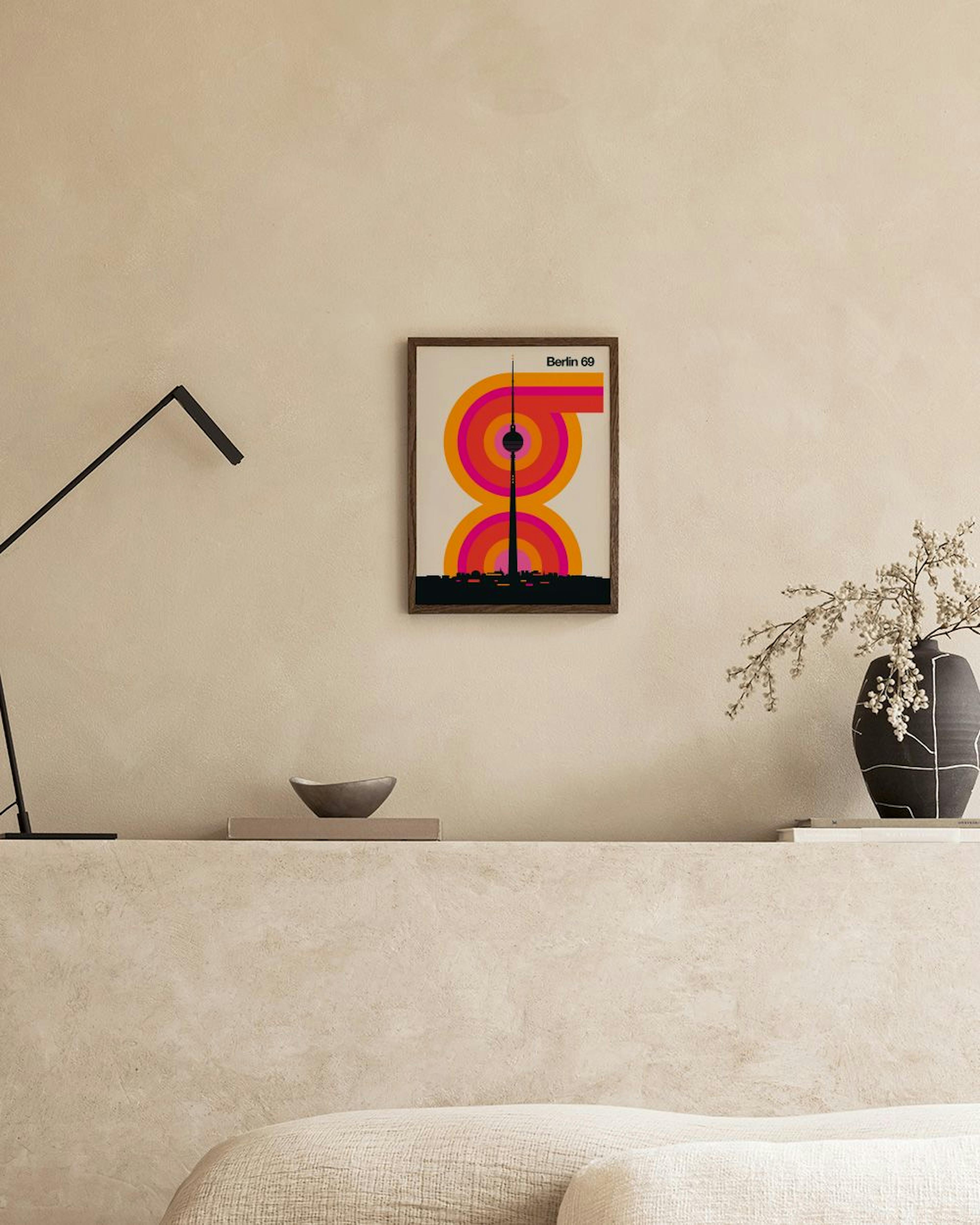 Poster Berlin 69 in Orange, Pink, Schwarz, Weiß, mit Fernsehturm und Stadtsilhouette, über einem Hocker mit Büchern.