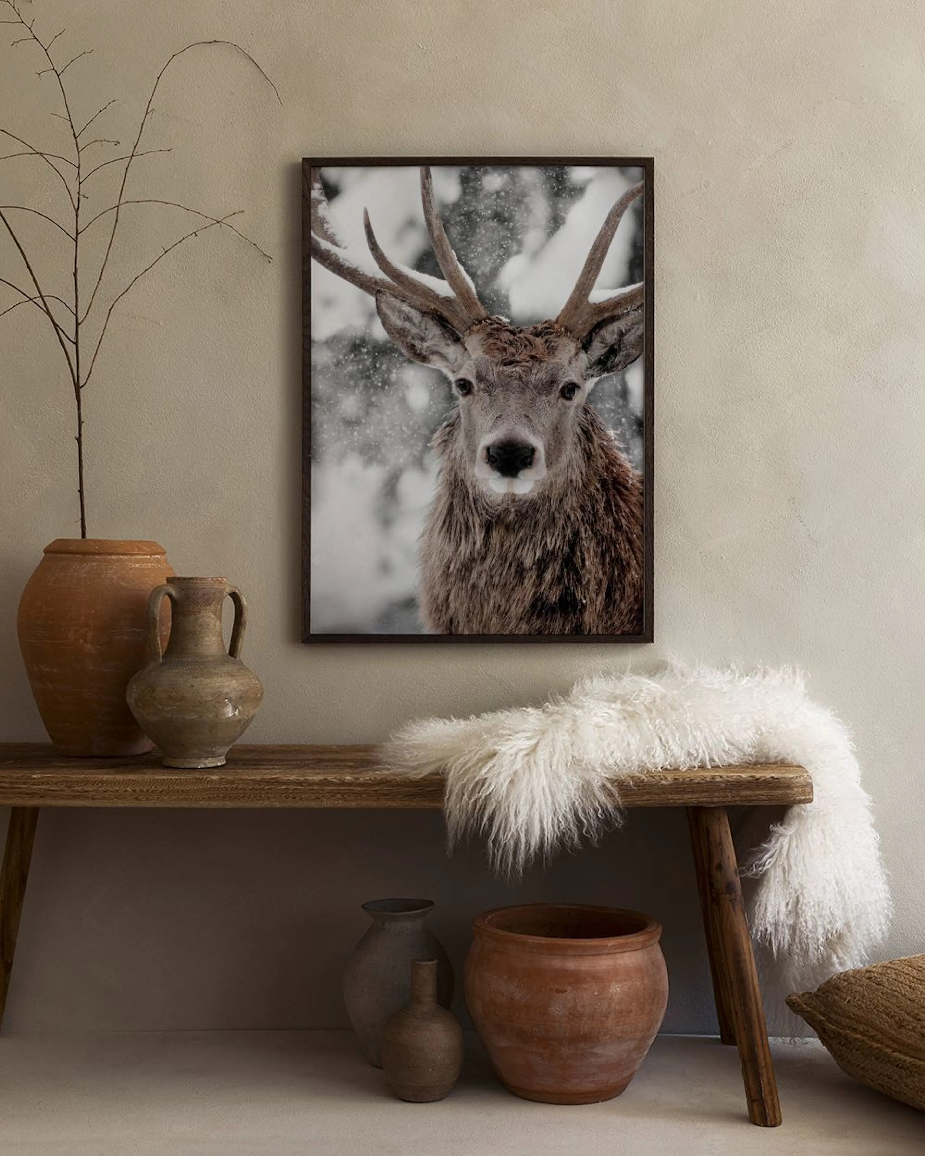 A print of a majestic deer with antlers in a snowy forest, hanging above a concrete bench with a book and vase.