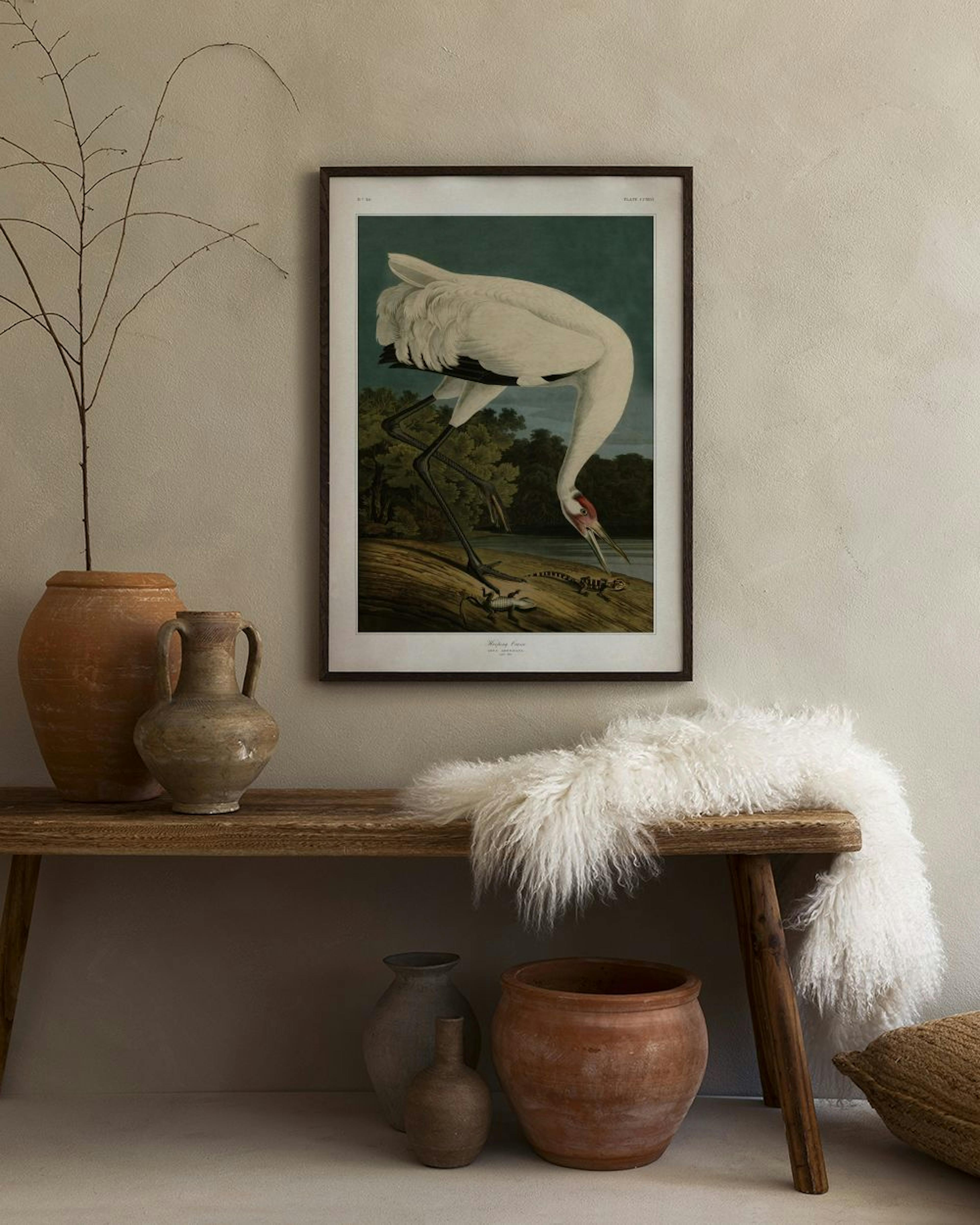 Poster med en trana som fångar ett djur vid en flod, hängandes i ett ljust rum ovanför en pall med böcker.