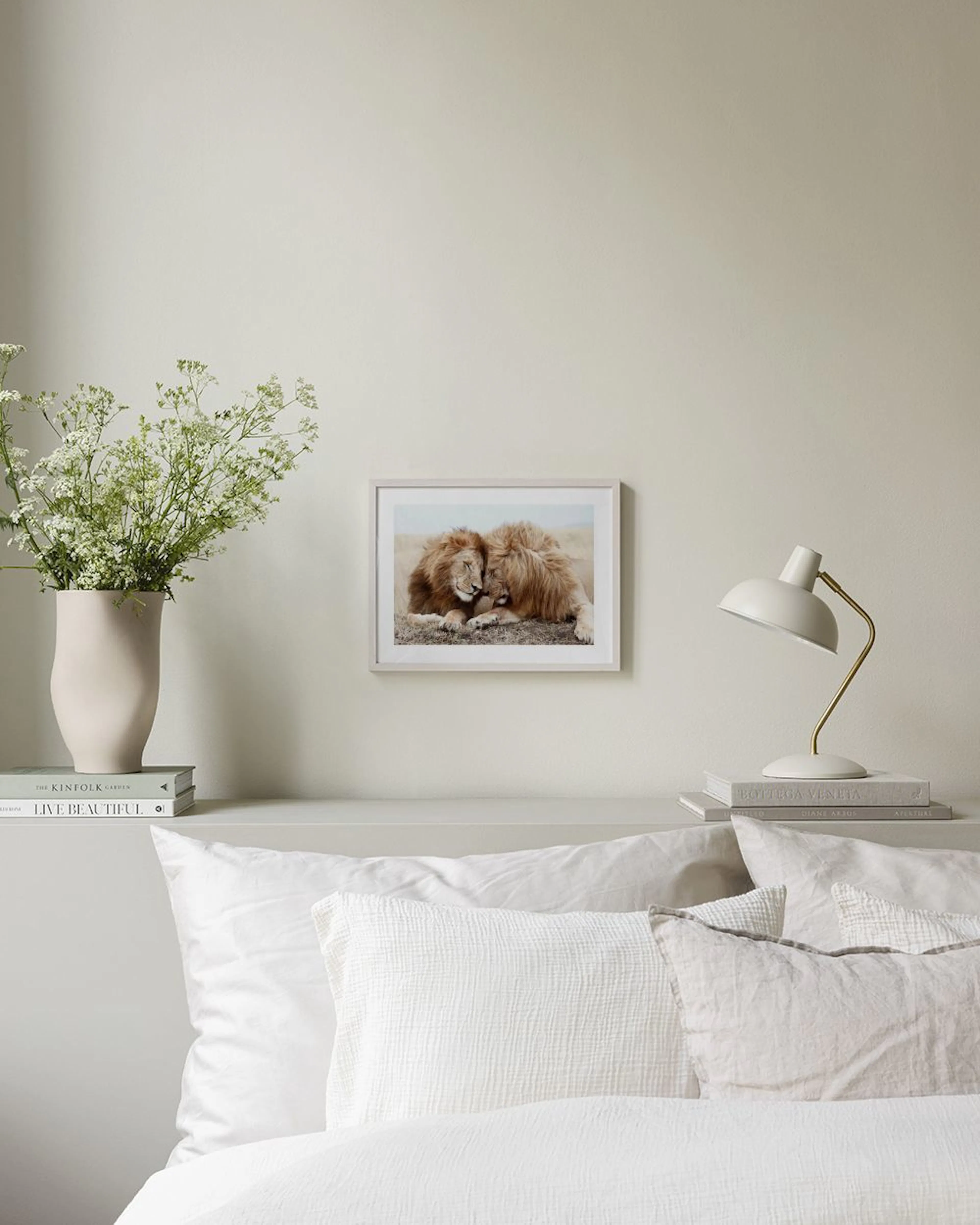 A print of two lions nuzzling, lying in dry grass, displayed on a wooden cabinet with a white lamp and vase.