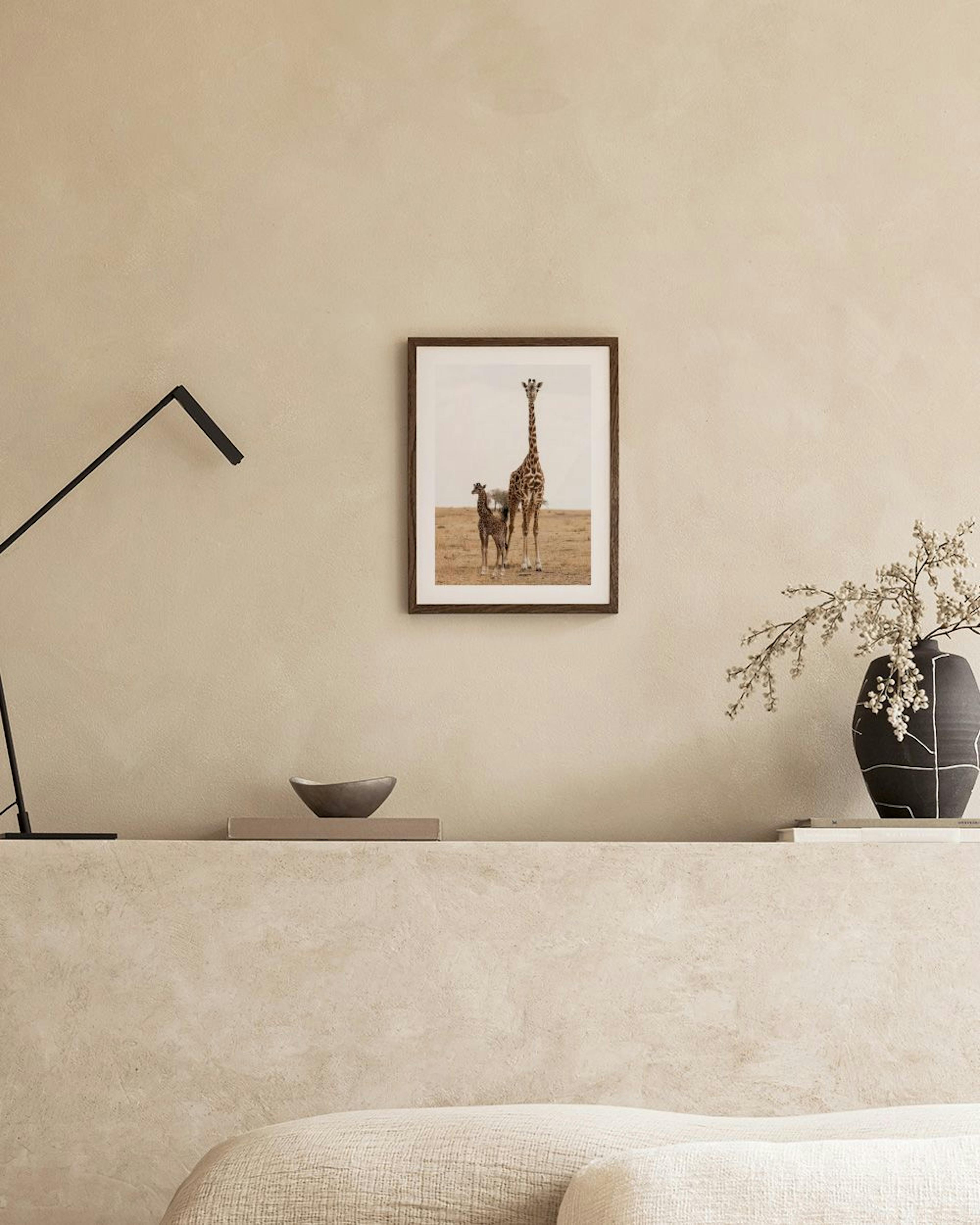 A print of a giraffe and its baby in a dry landscape, displayed above a light wood desk with a cactus and lamp.